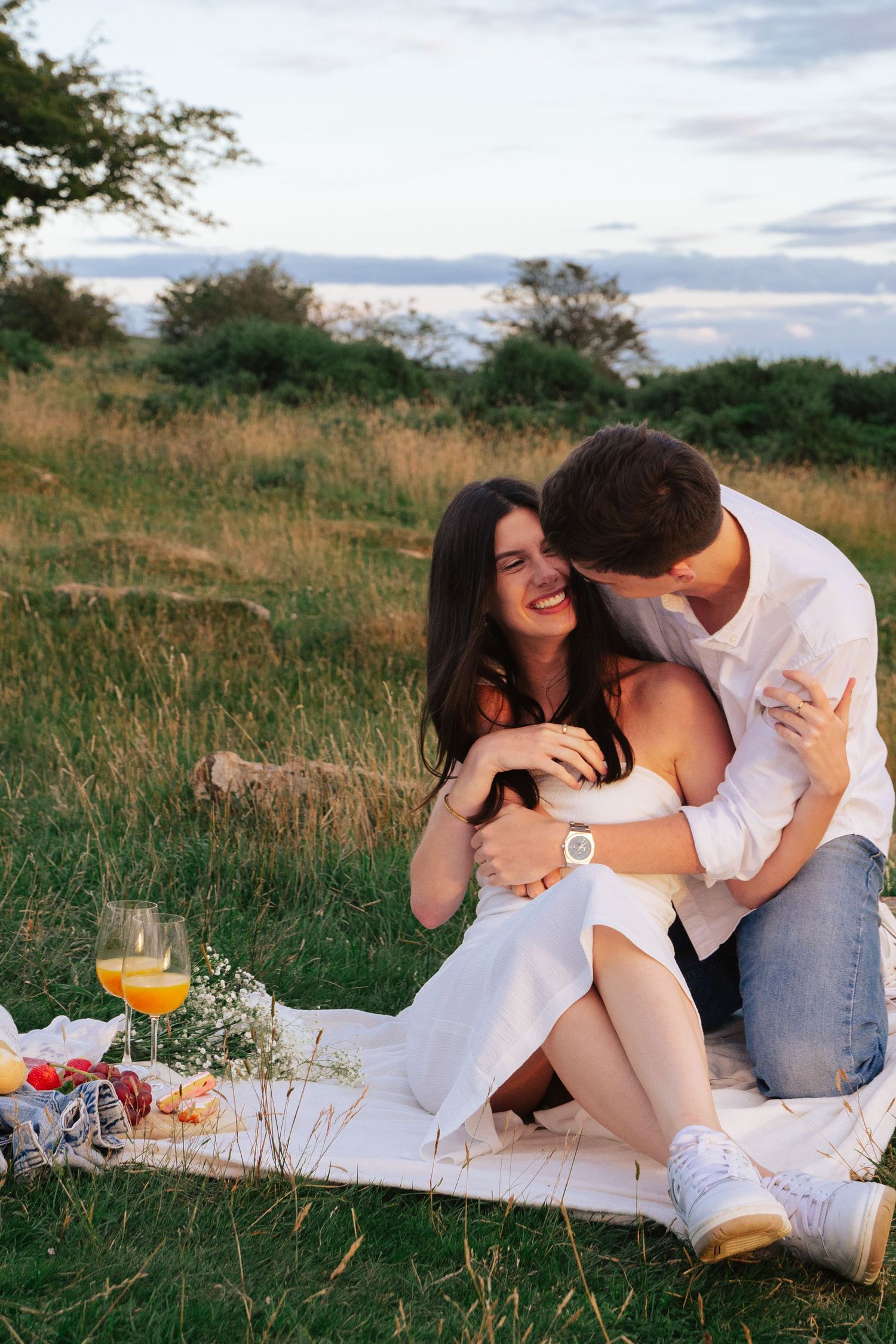 Couple having a romantic picnic on a white blanket in a grassy field at sunset.