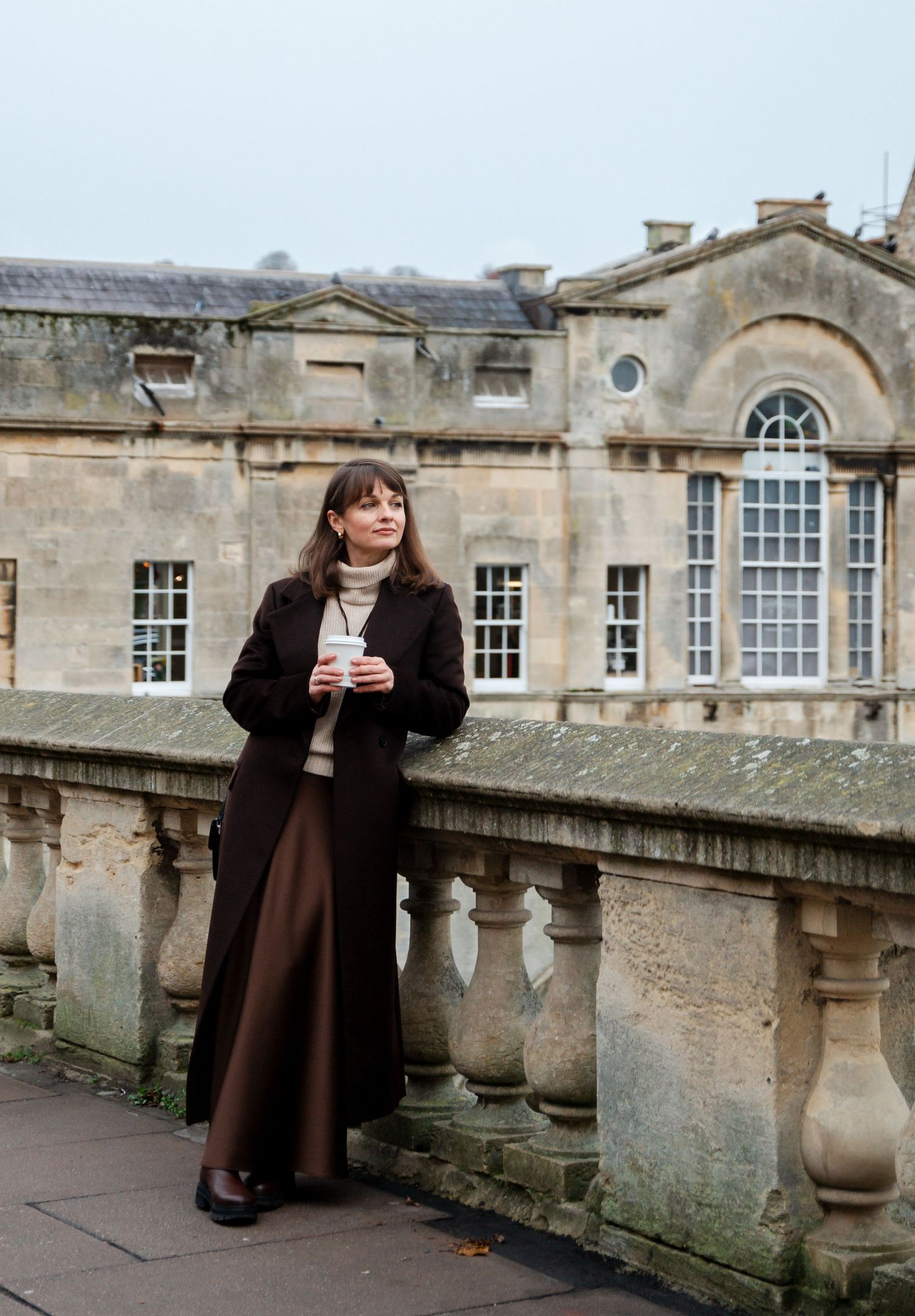 Figure in long brown coat standing against historic stone balustrade and building facade.