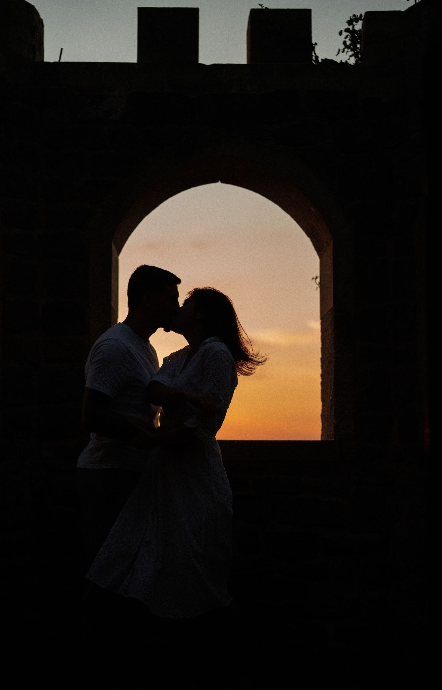 Silhouetted figures embracing in an arched window during a dramatic sunset.