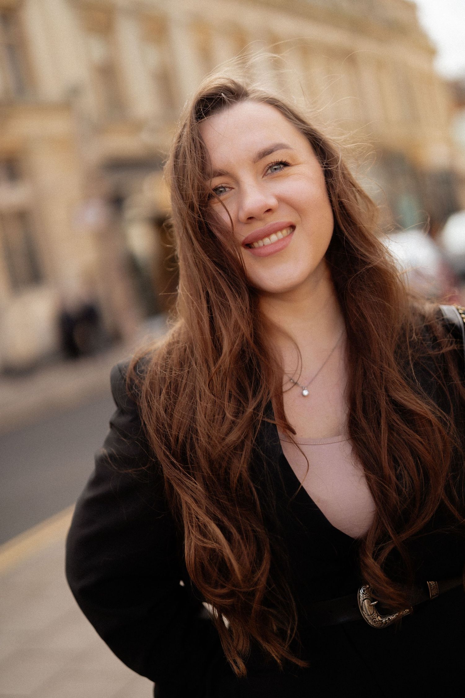 Smiling young woman with long brown hair wearing a black jacket, posing on a city street.