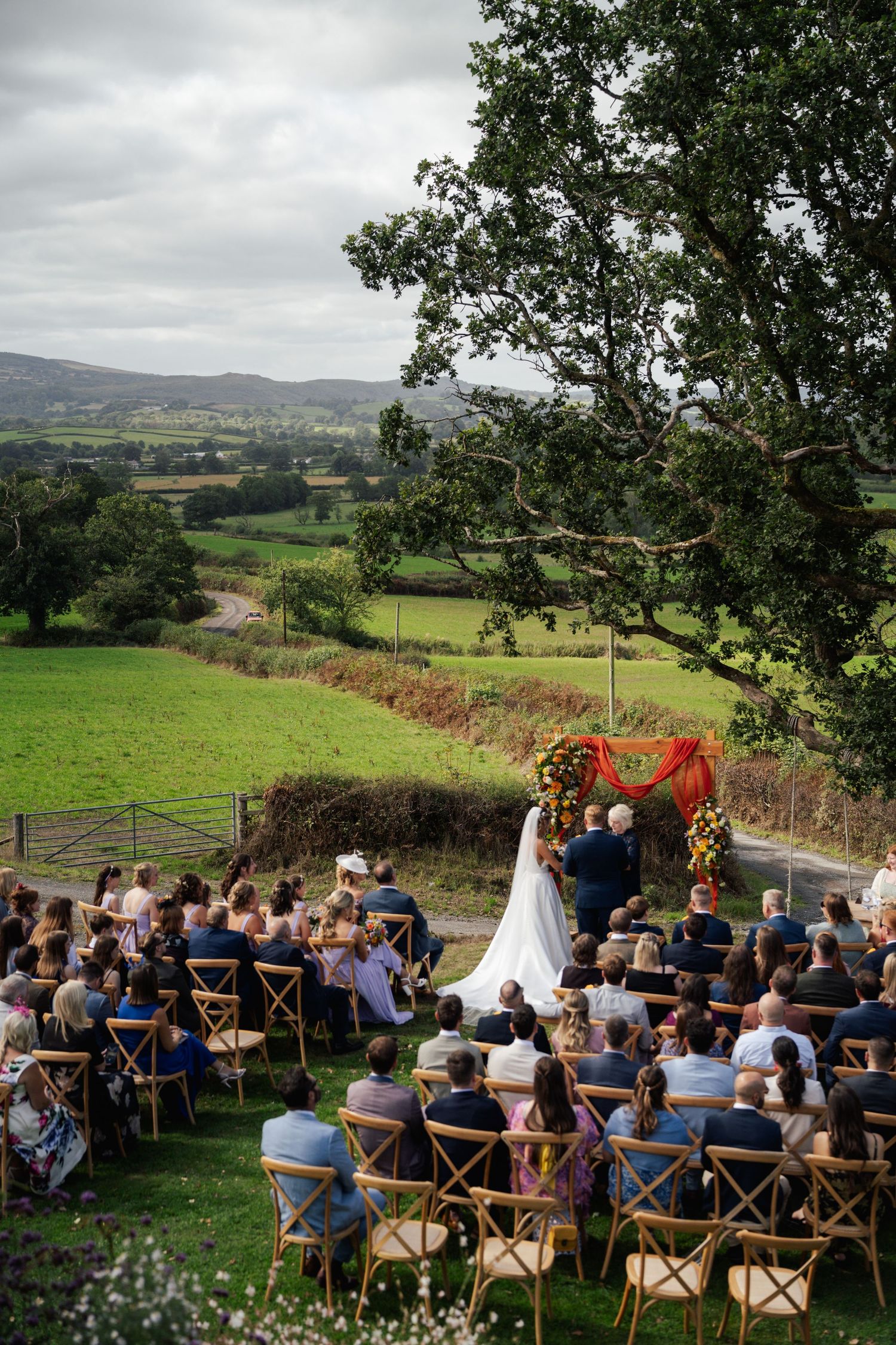 Outdoor wedding ceremony with guests seated in wooden chairs overlooking a scenic green valley and rolling hills.