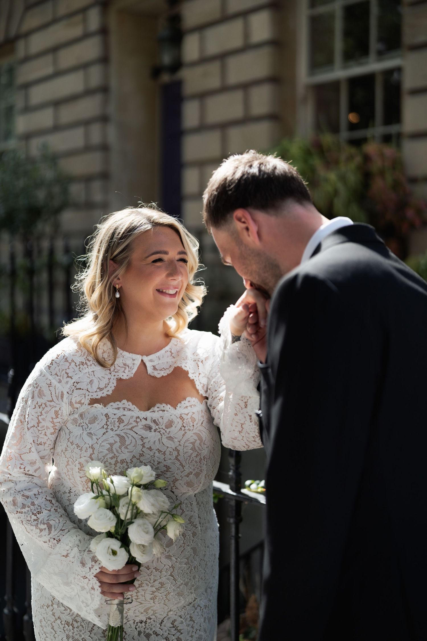 Bride in lace dress holding white bouquet smiles at groom in dark suit outdoors on wedding day.
