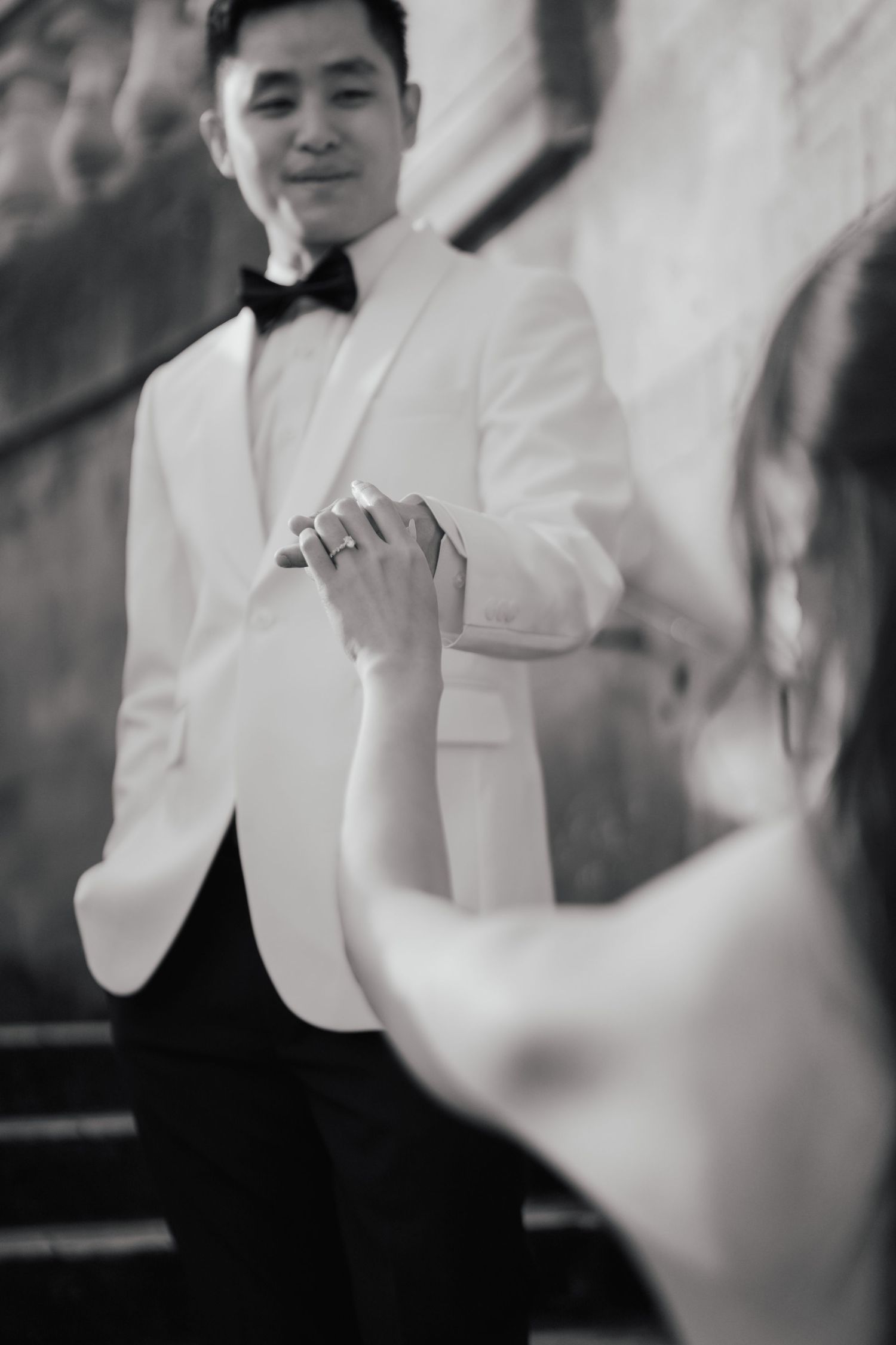 Black and white photo of groom in white tuxedo holding hands with bride during romantic wedding dance.