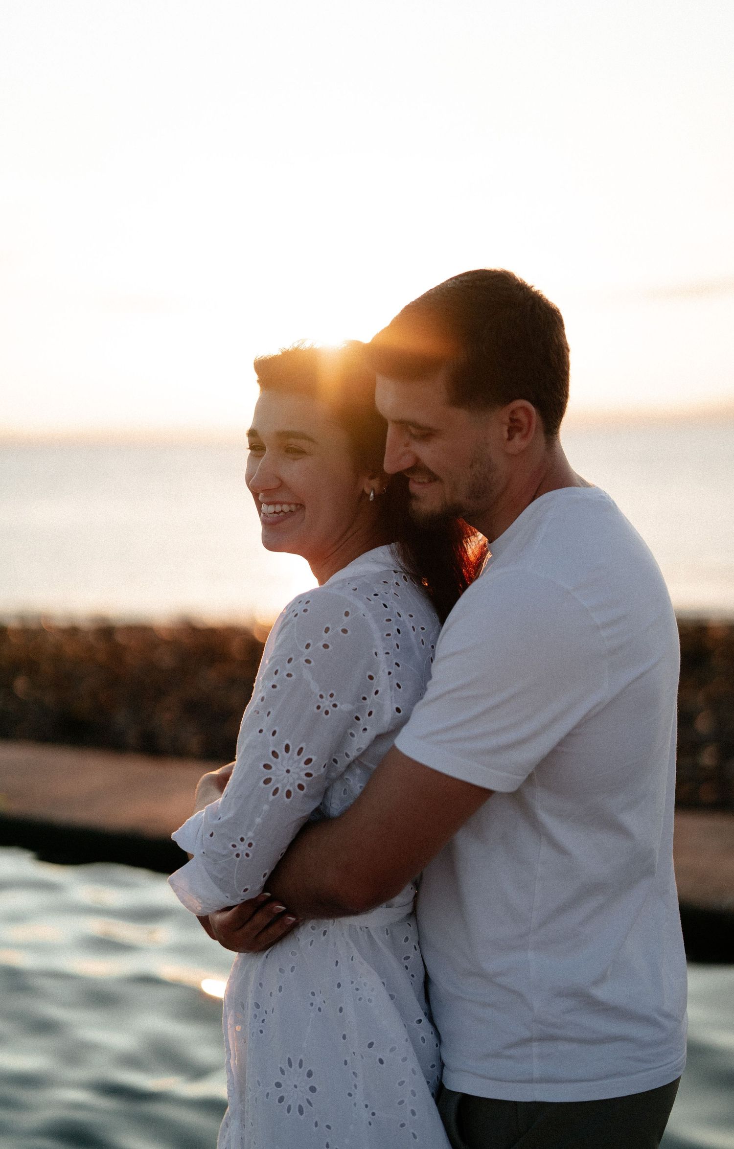 Couple embracing at sunset by the ocean, woman in white dress laughing joyfully as man holds her from behind.