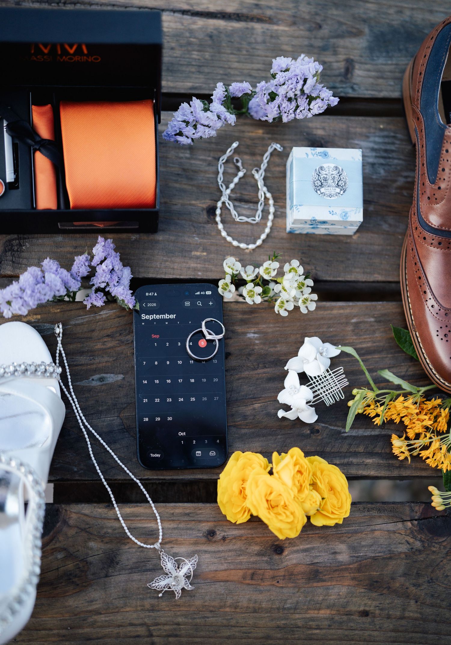 Flat lay of wedding accessories including orange tie, brown shoes, jewelry, yellow roses, and a phone showing September calendar on wood.