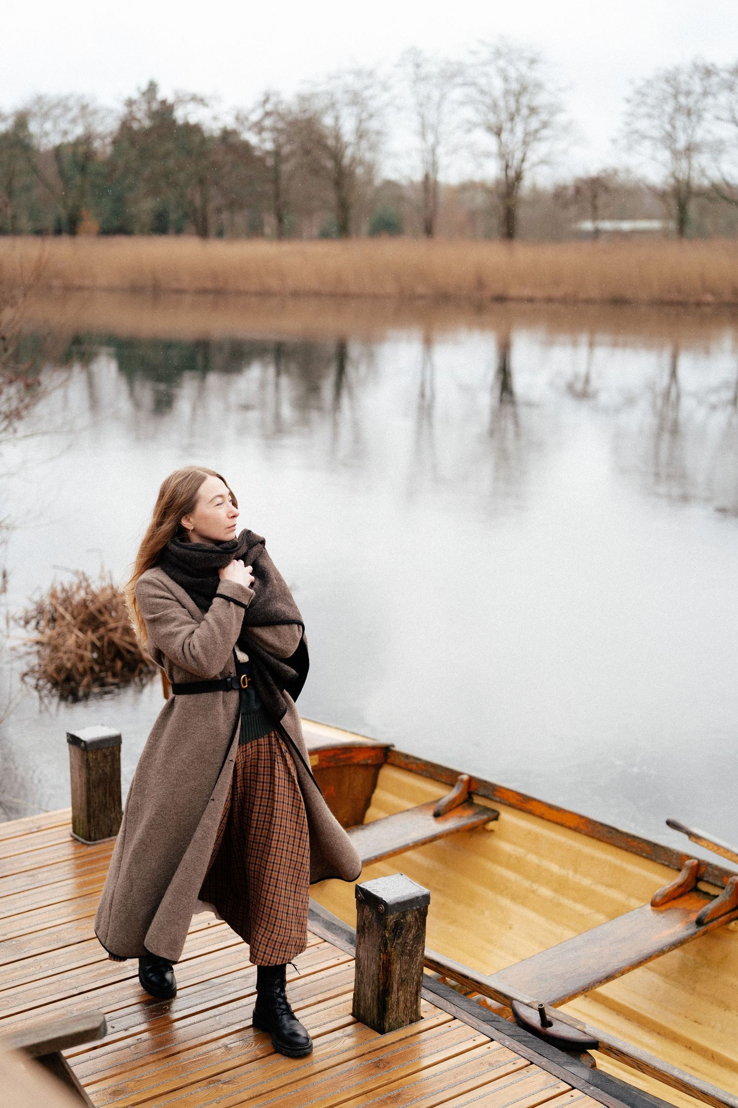 A person sits on a wooden dock by a serene lake on a winter day wearing a brown coat and plaid scarf.