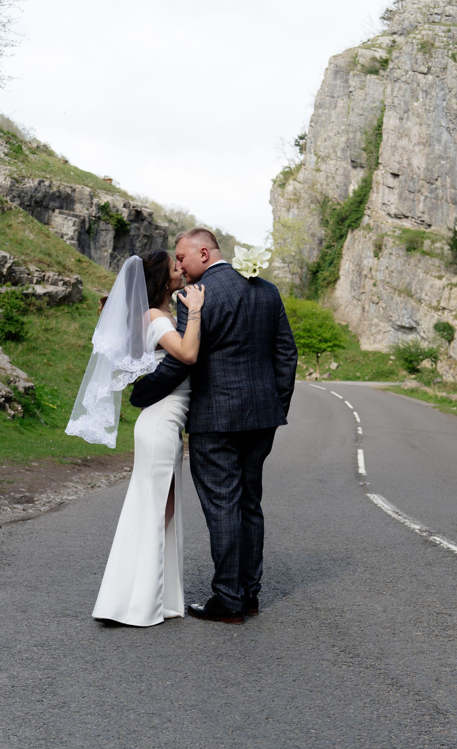 A couple shares a romantic moment on a winding mountain road with tall rock formations in the background.