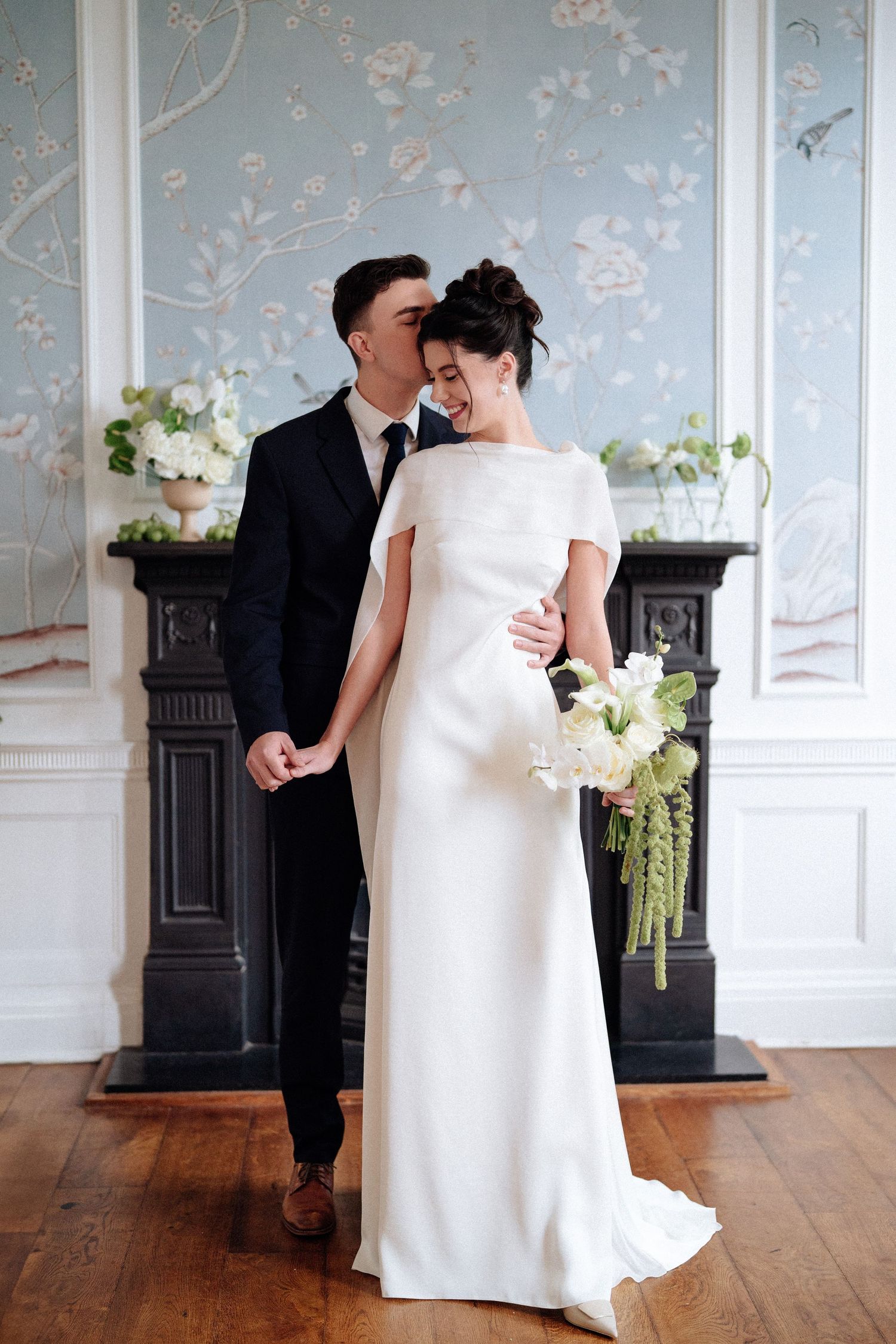 A romantic wedding couple embraces in an elegant room with blue floral wallpaper and hardwood floors.