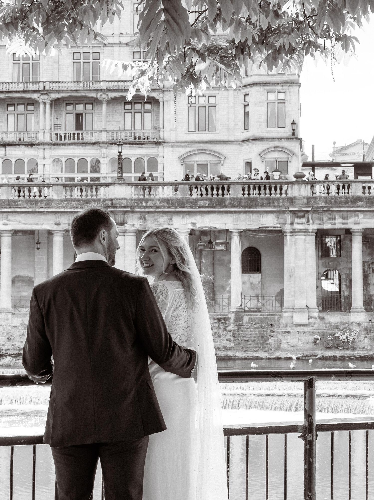Black and white photo of a bride and groom standing by a railing, gazing at a grand historic building.