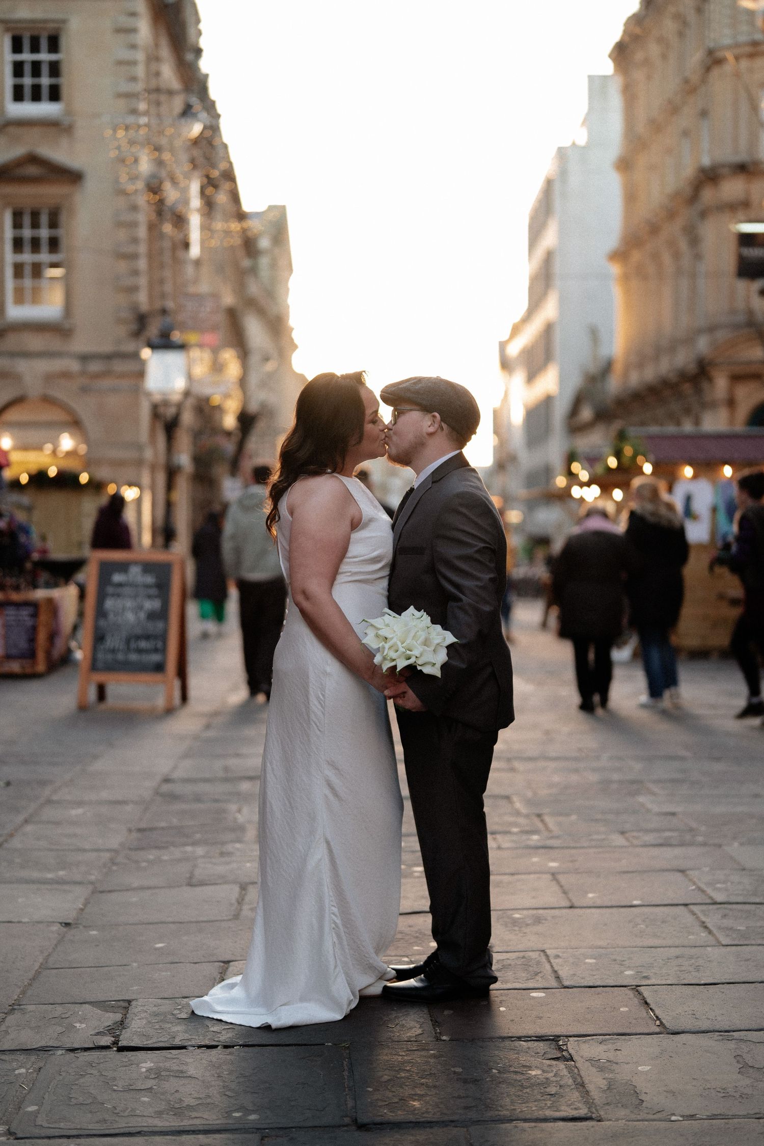 A couple in wedding attire share romantic moments on a historic cobblestone street at sunset in Bath, England.
