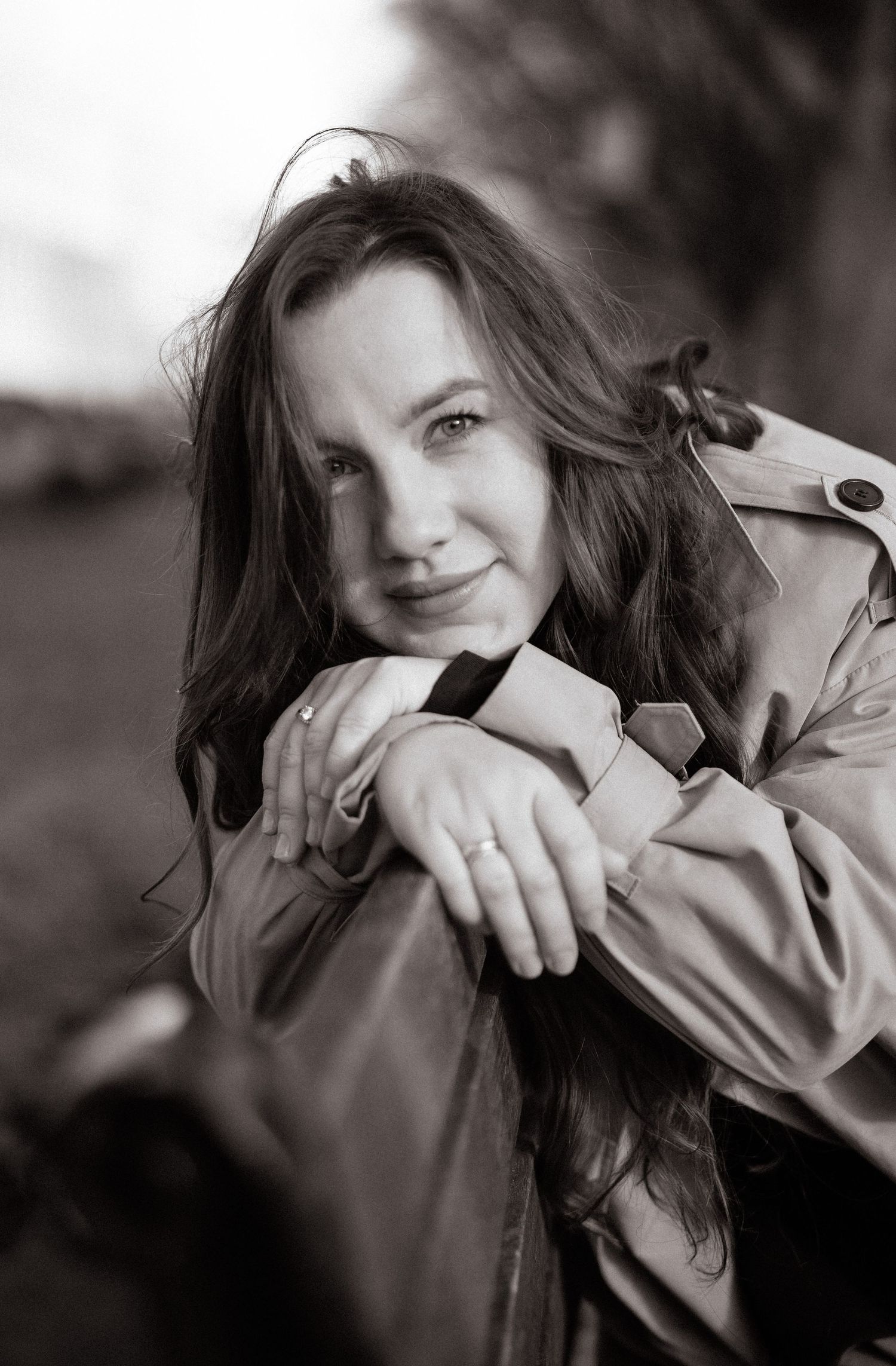 Black and white portrait of a smiling young woman with long hair, wearing a trench coat, resting her arms on a railing.