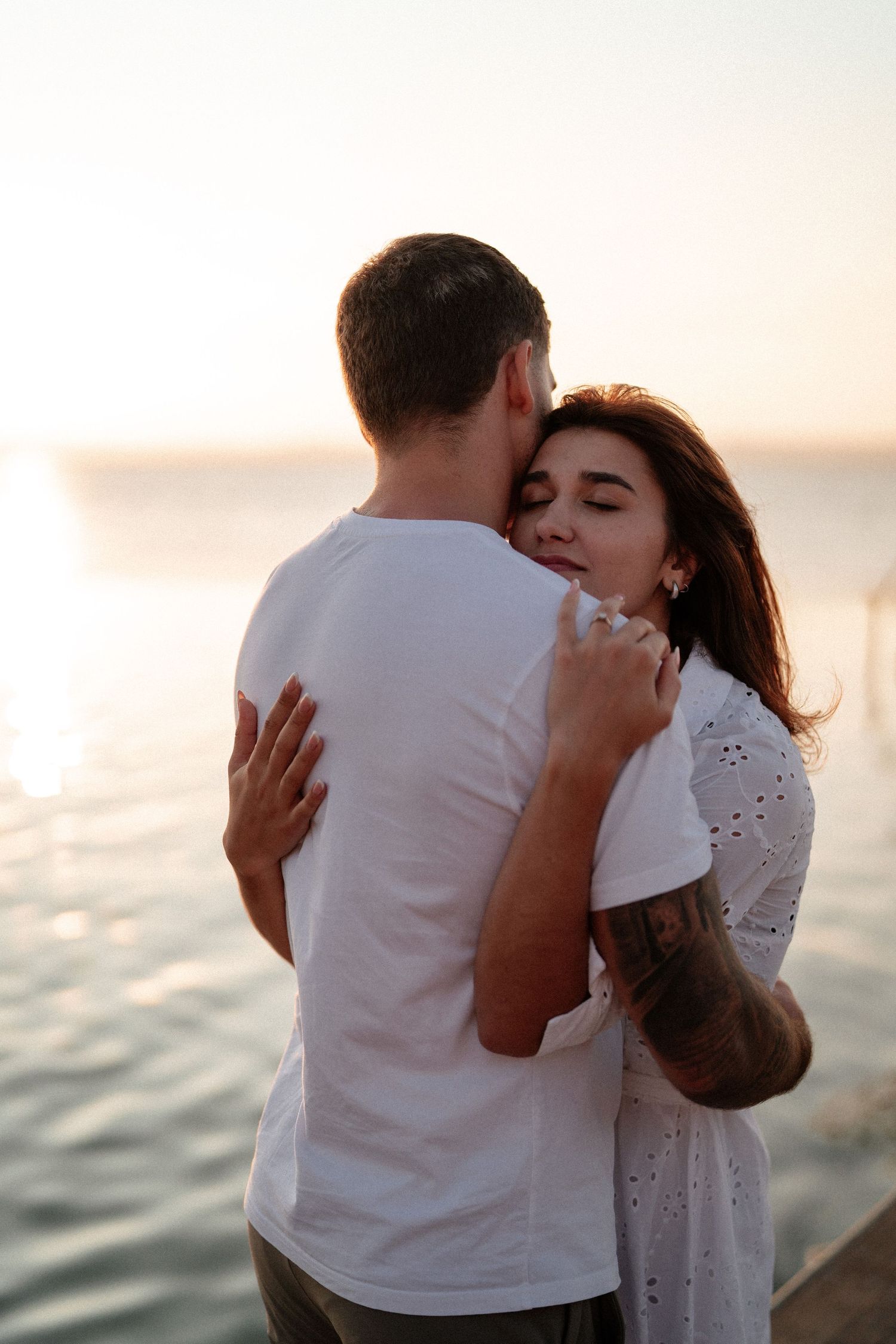 Couple embracing romantically at sunset by the ocean, woman in white dress with tattoo, golden hour backdrop.