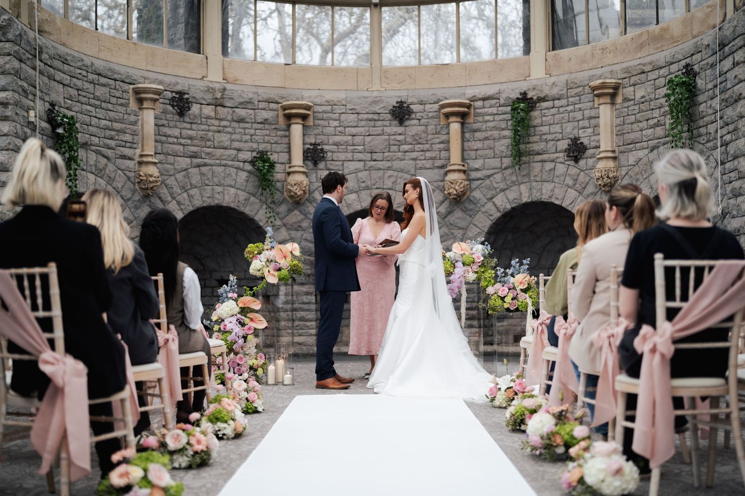 Wedding party in formal attire gathers inside elegant stone rotunda with arched windows and floral decorations.