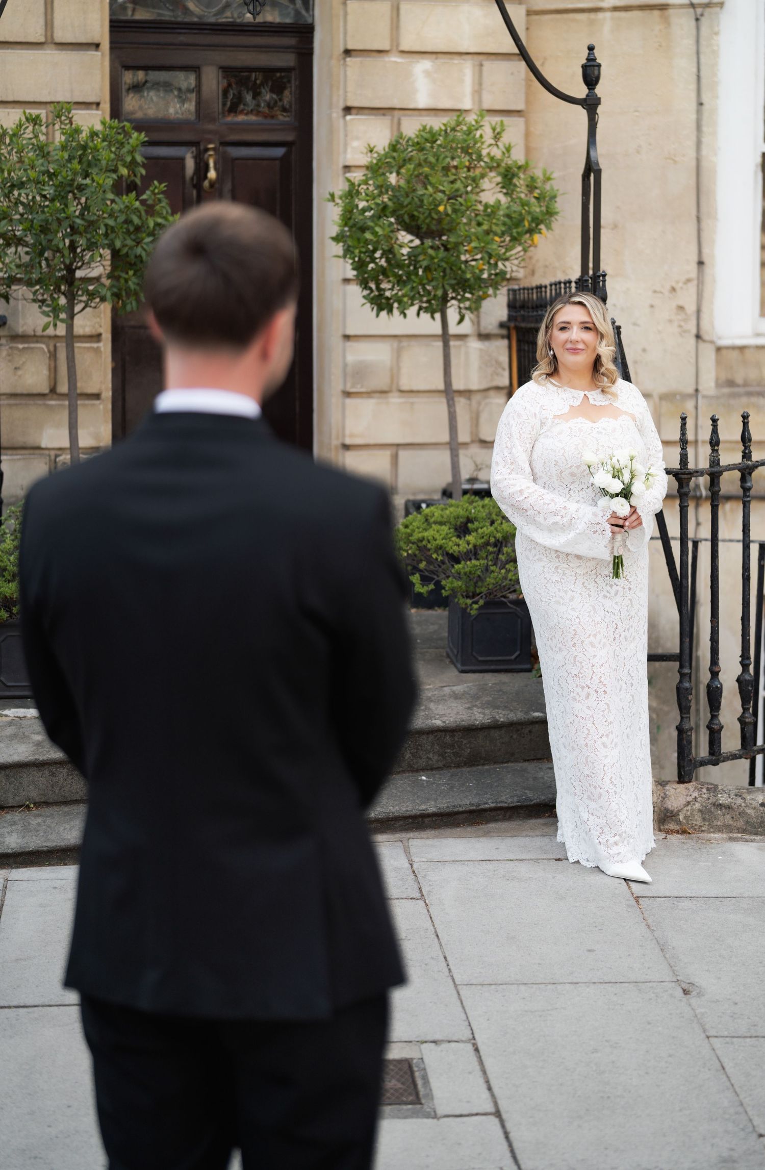 Bride in white lace gown holds bouquet as groom in black suit faces her outside a stone building.