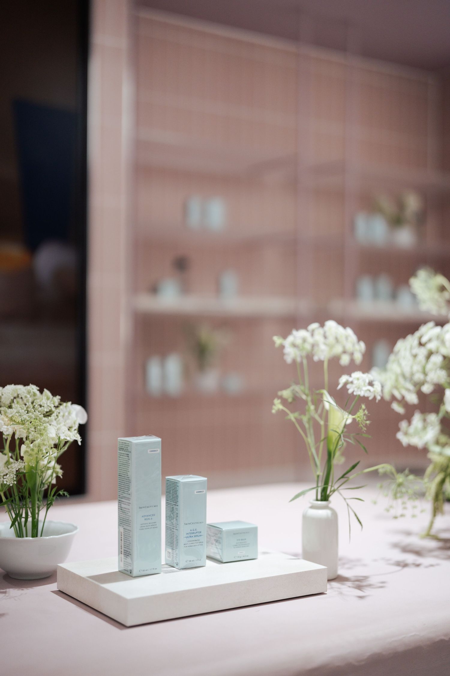 Elegant skincare product display with light blue boxes on white tray, surrounded by white wildflowers in a pink-toned spa setting.