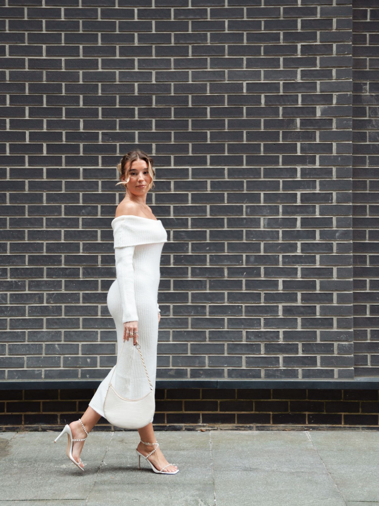 Woman in flowing white dress poses gracefully against dark brick wall.