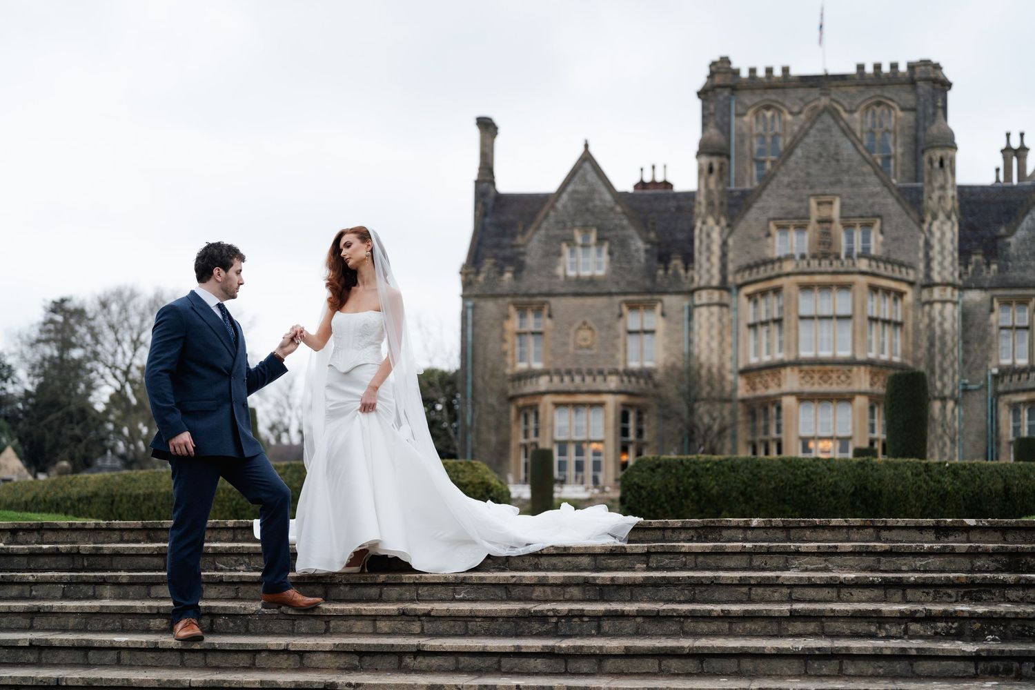 A couple sits on stone steps outside a grand historic manor house in a romantic wedding photo.