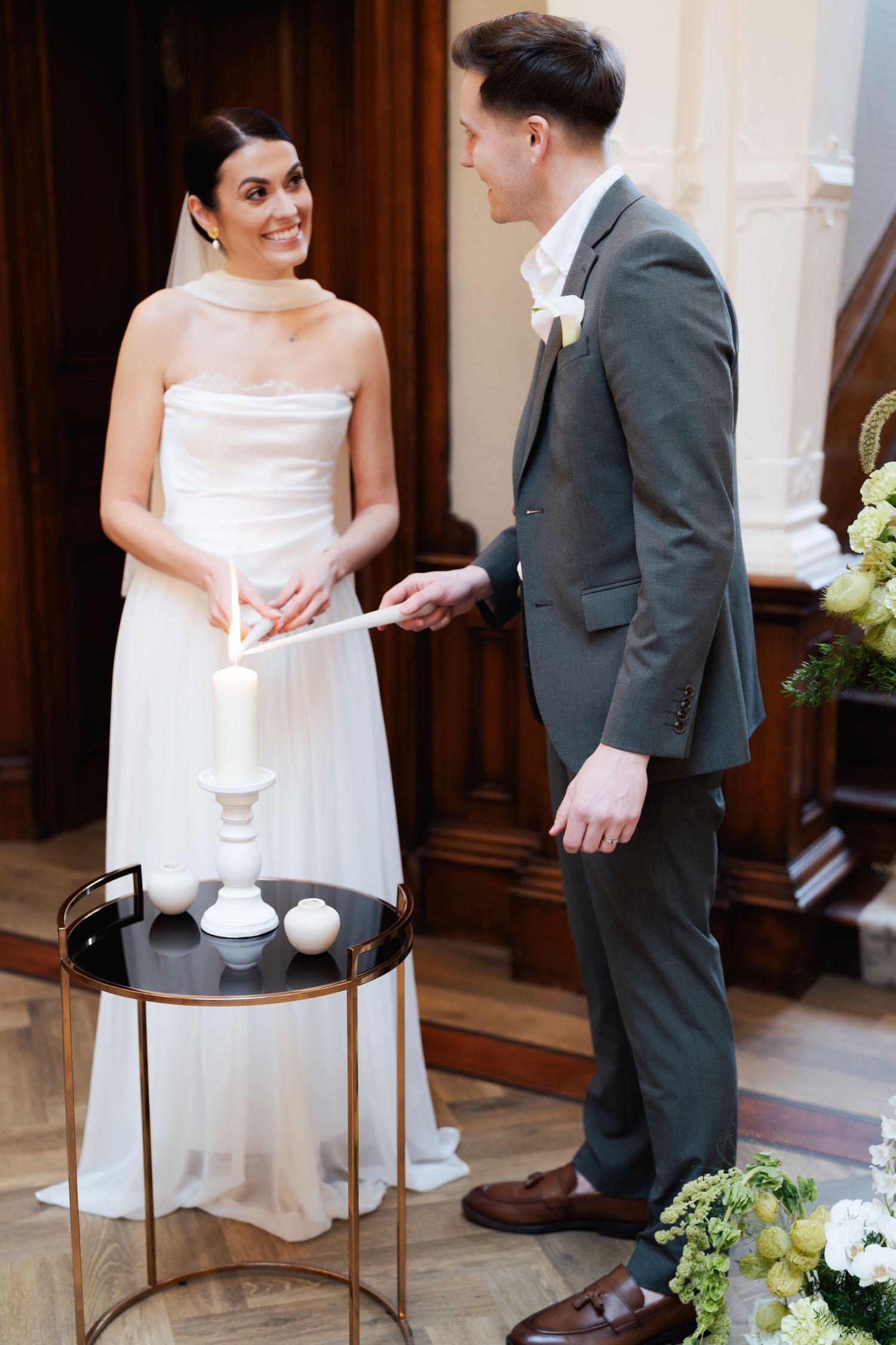 A bride in a white strapless gown and groom in gray suit stand by a small table with candles during their wedding ceremony.