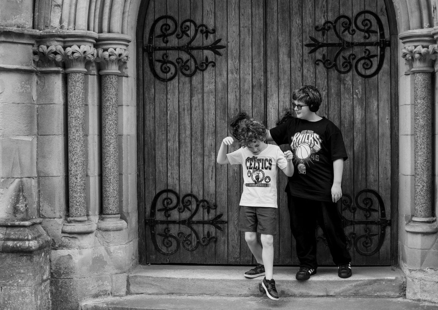 Two children stand in front of an ornate gothic doorway with decorative iron scrollwork and stone columns.