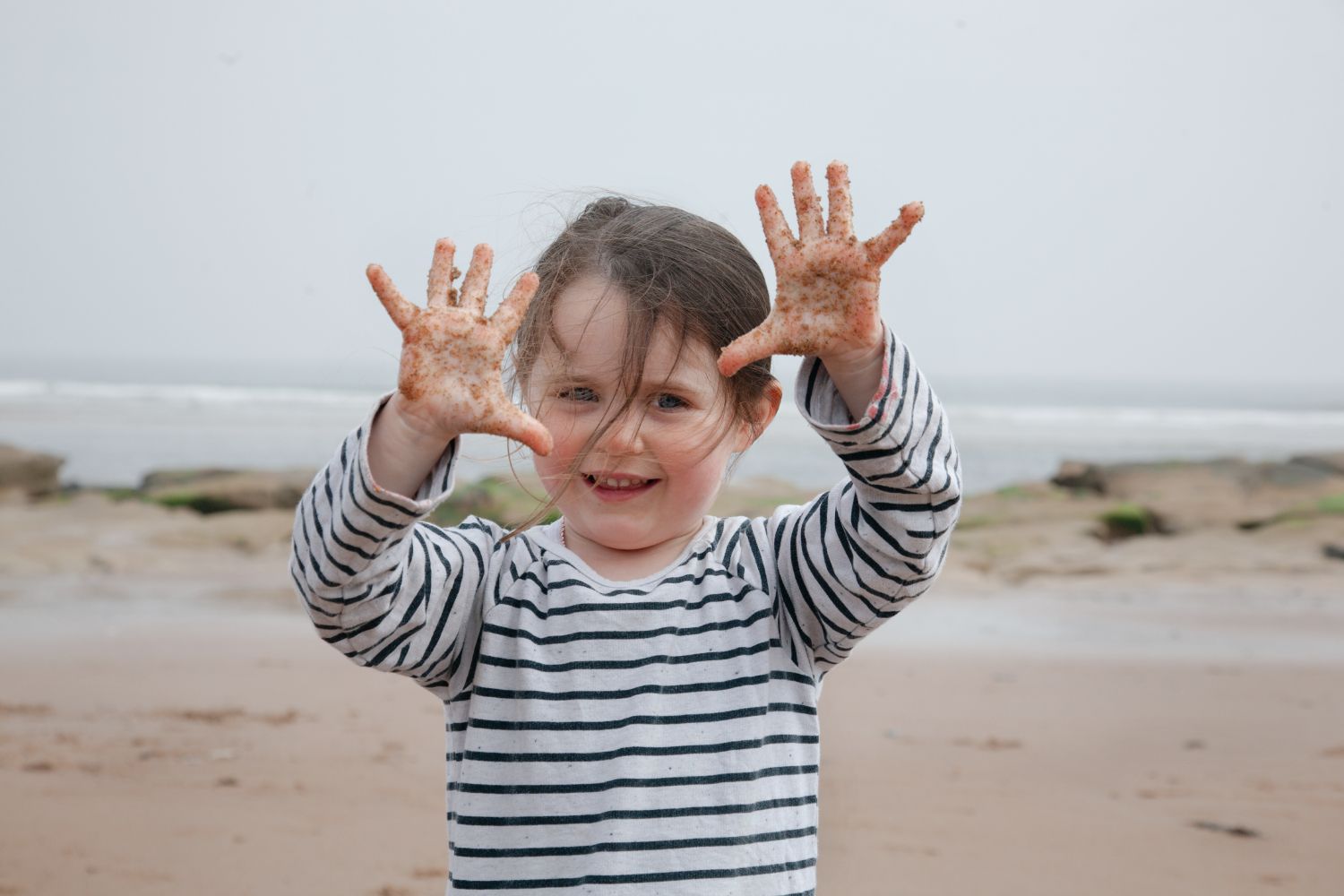 Natural portrait of a child playing outdoors on a baech in a striped shirt showing sand covered hands.