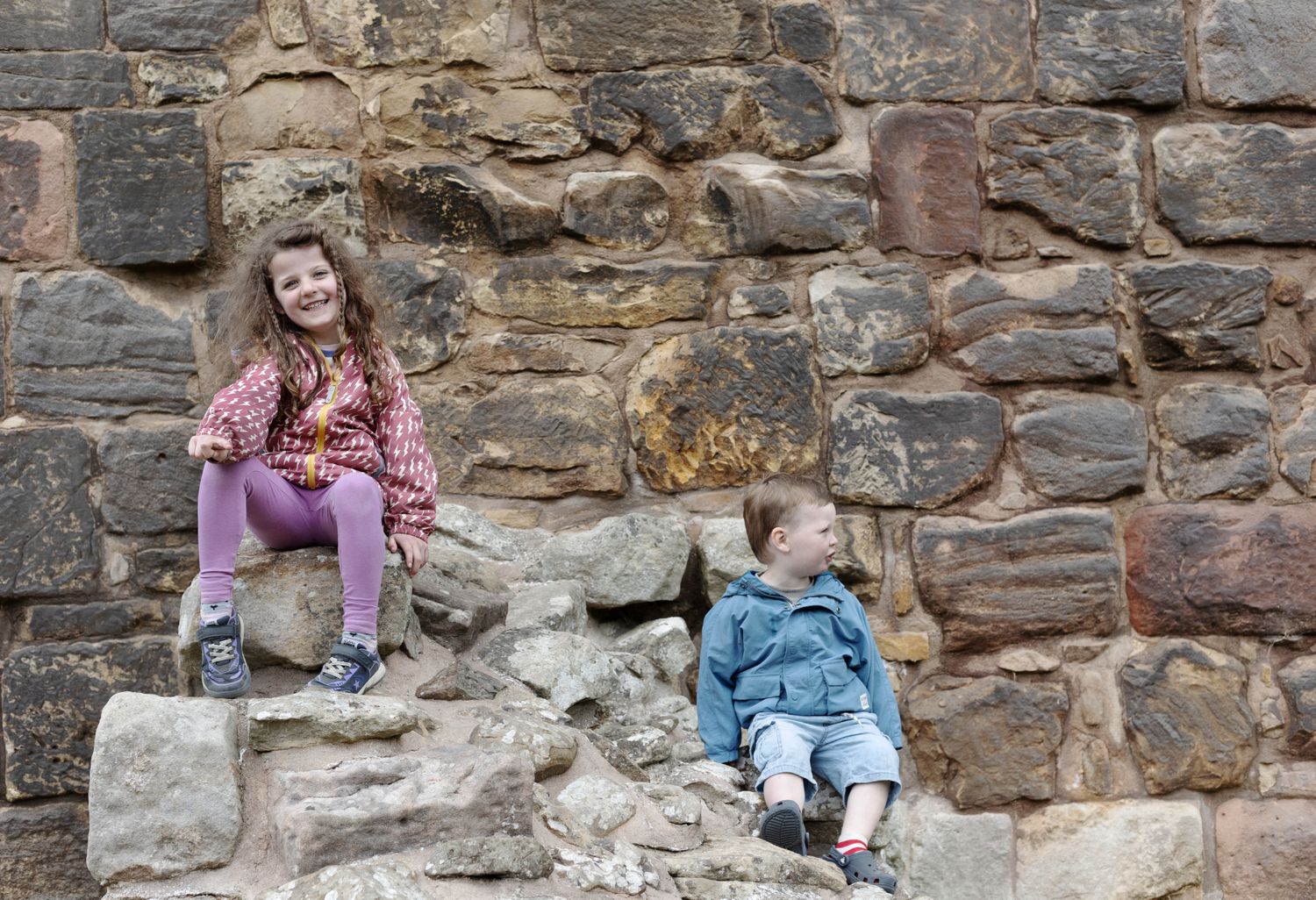 Two children in relaxed poses perched on a stone ruin with a rustic wall behind.