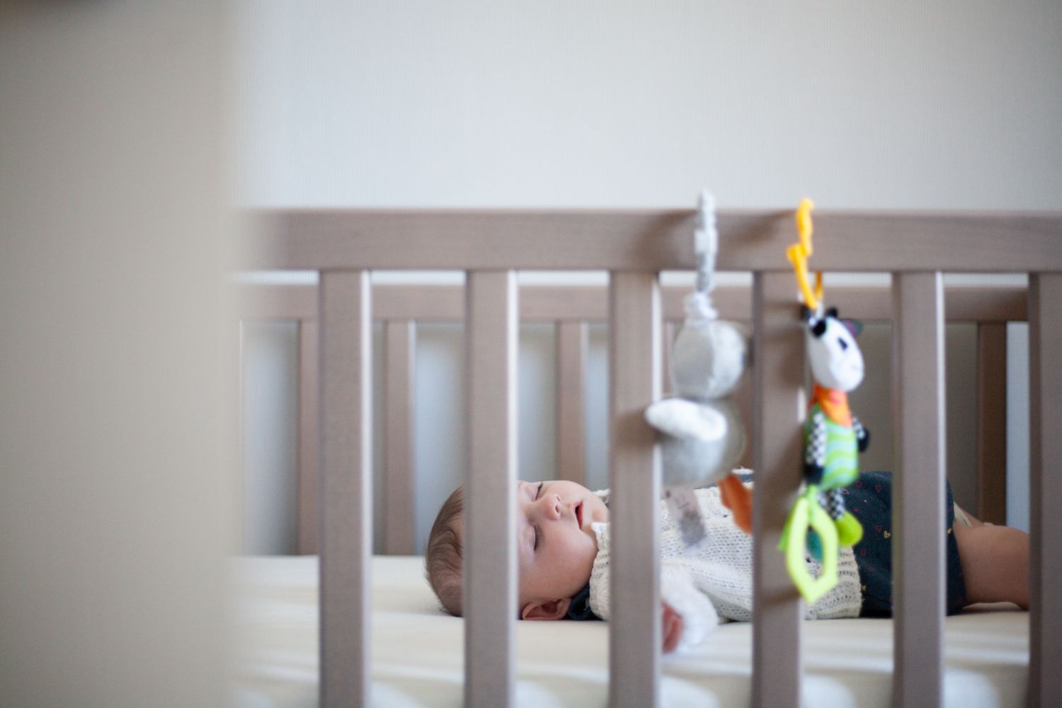 Mobile toys hanging from cot frame bars in a nursery with a sleeping toddler