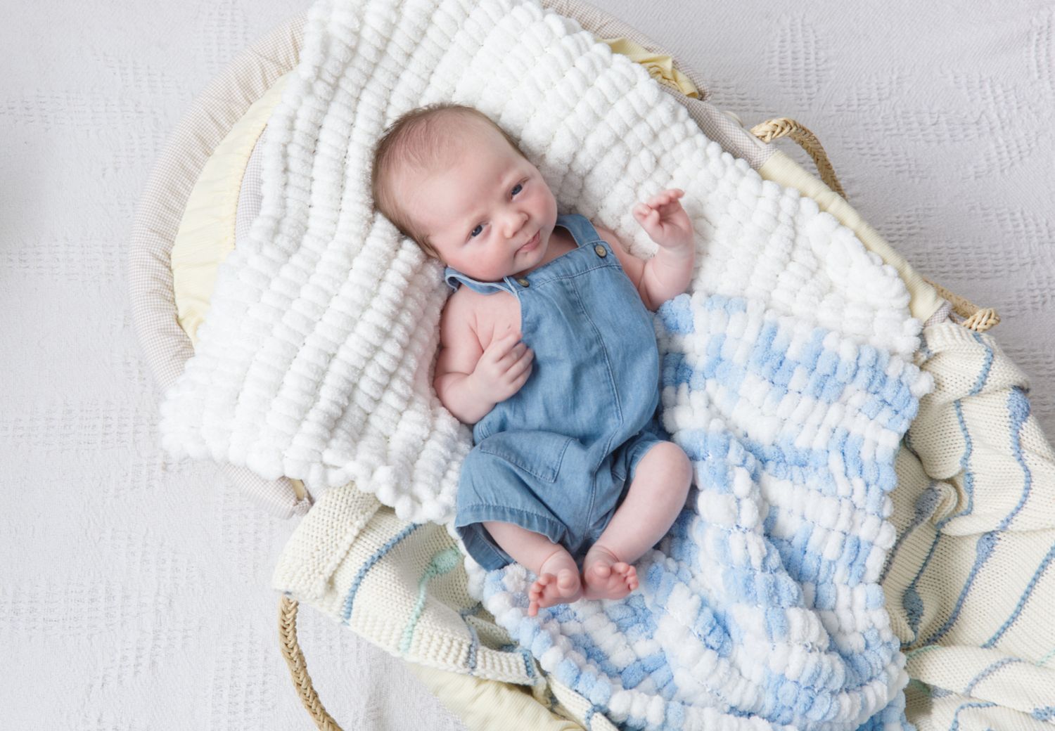 Newborn baby in blue denim romper lying in cozy woven basket with soft white and blue blankets.
