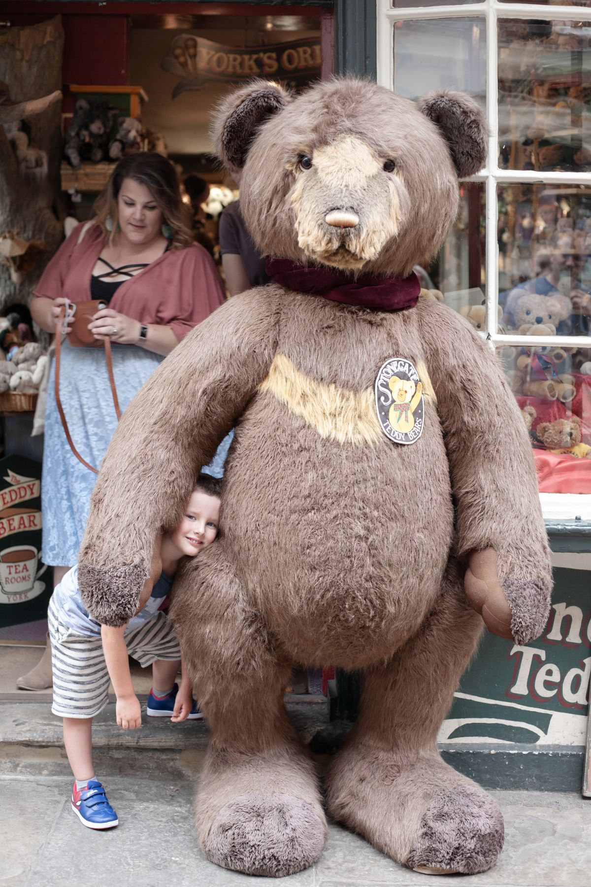 Location family portrait of a child peeping out from behind a very large teddy bear at a storefront with their parent close by