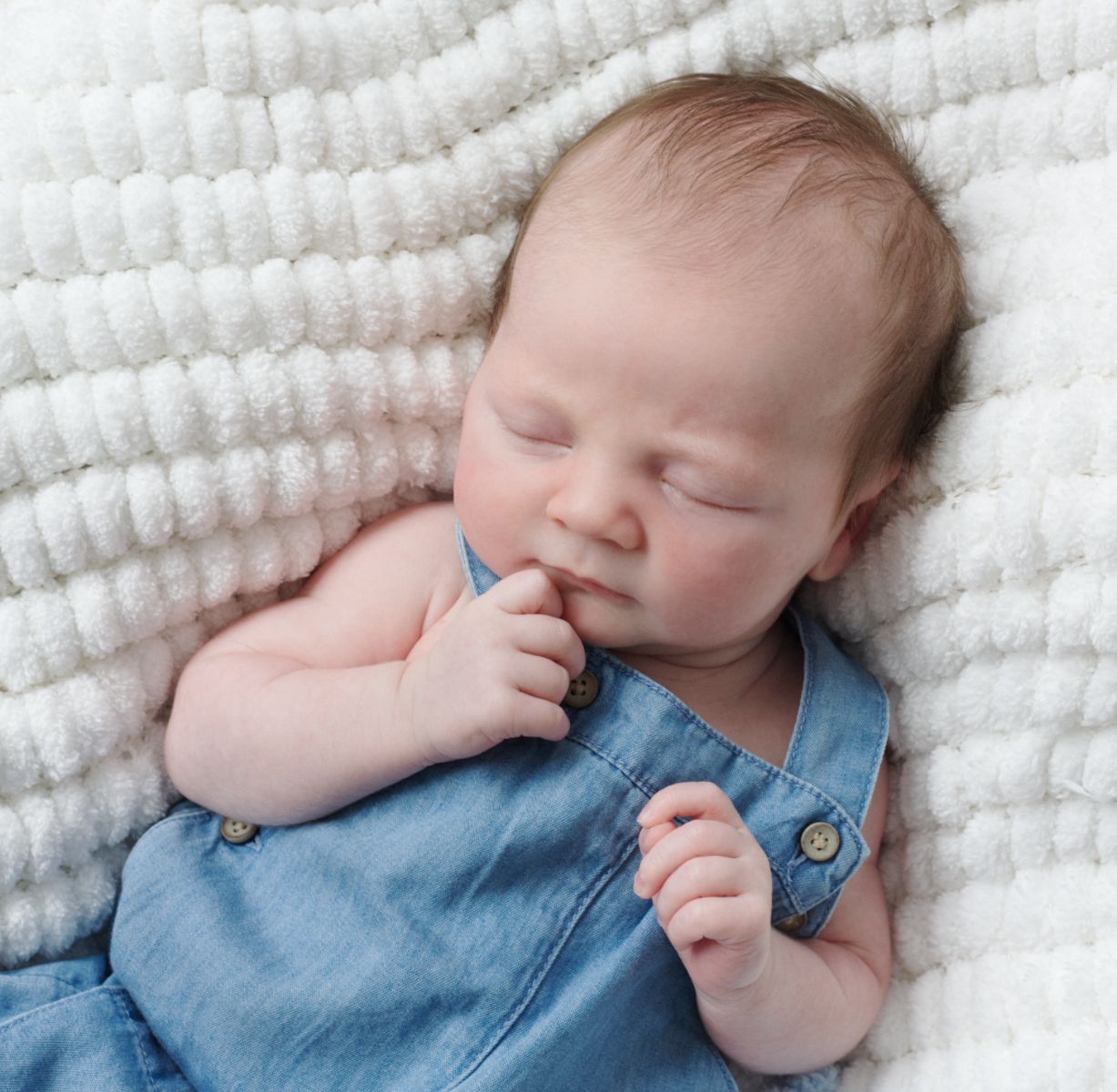 Sleeping baby wearing denim overalls rests peacefully on textured white blanket.