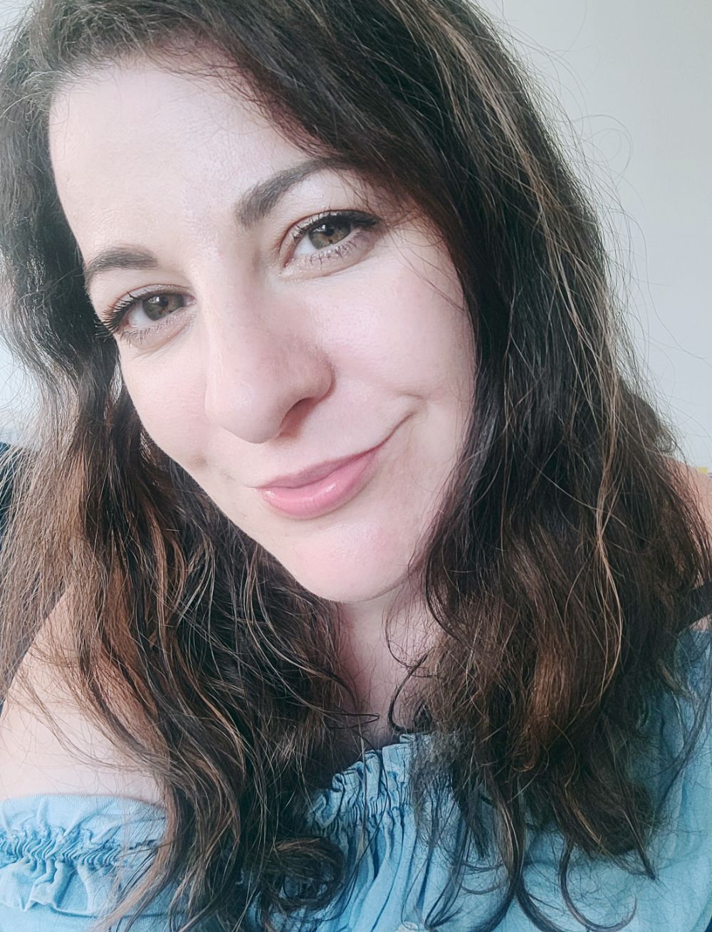 A person with wavy dark hair and a gentle smile looks at camera while wearing a light blue ruffled top.