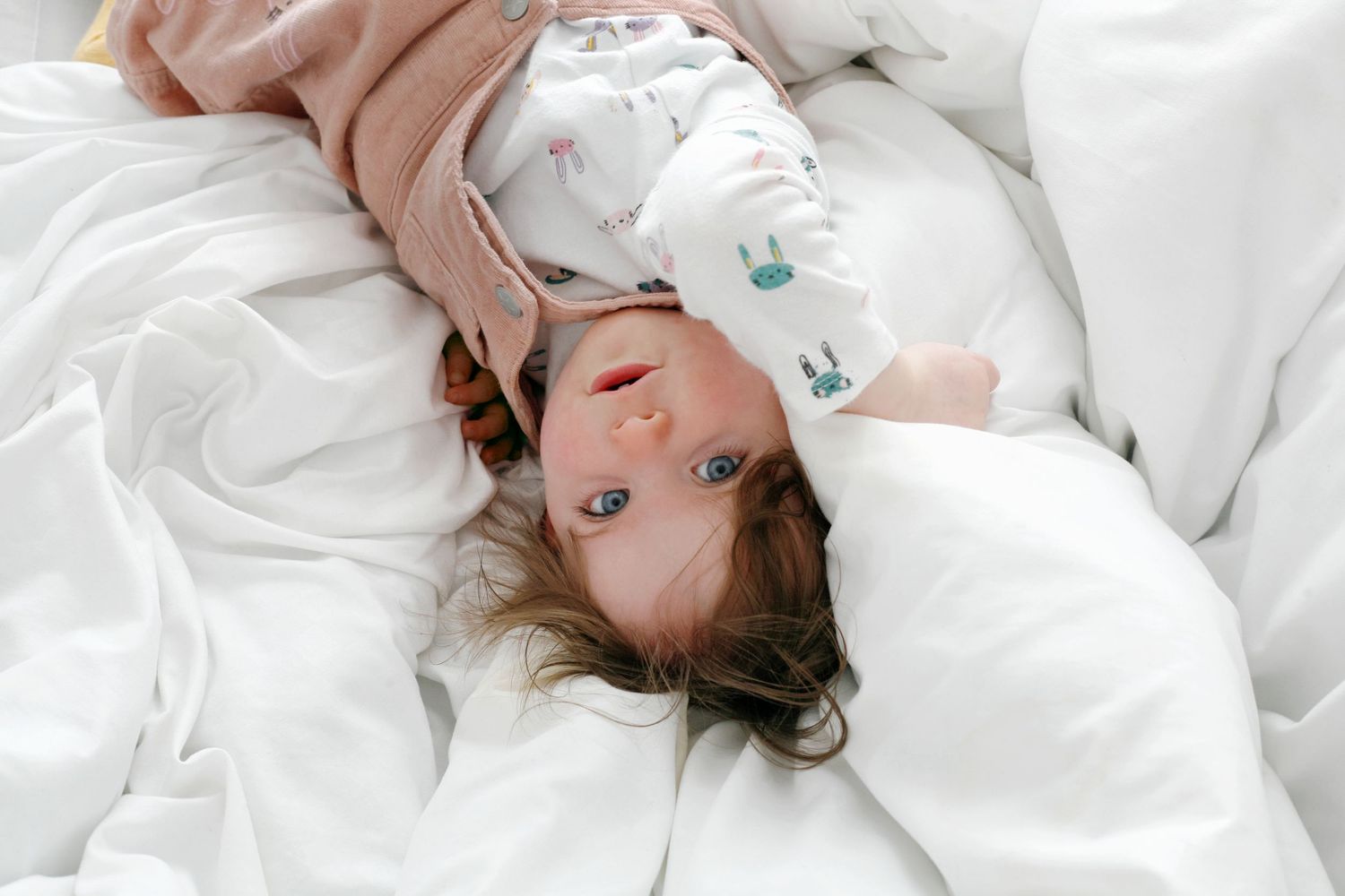 A child in pink dungarees, captured in a natural moment on a soft white duvet