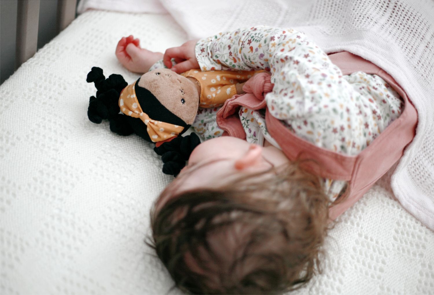 A small child rests on a white blanket wearing pink dungarees while holding a plush stuffed toy.