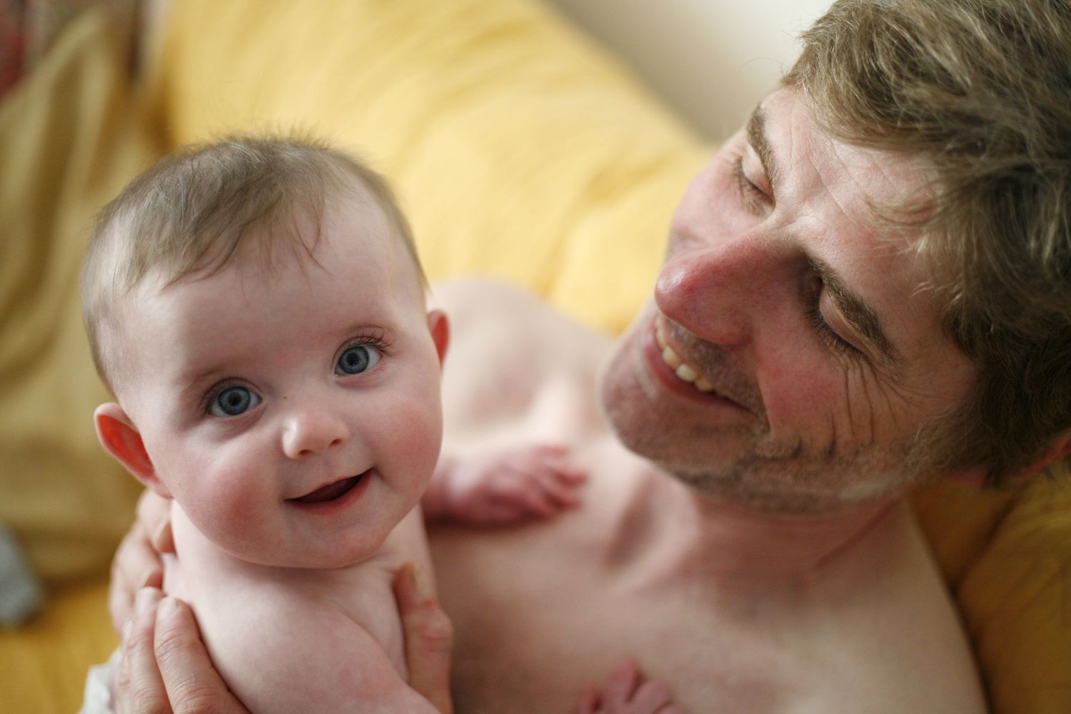 A happy baby and adult share a tender moment while relaxing on a yellow coloured couch. Both are smiling