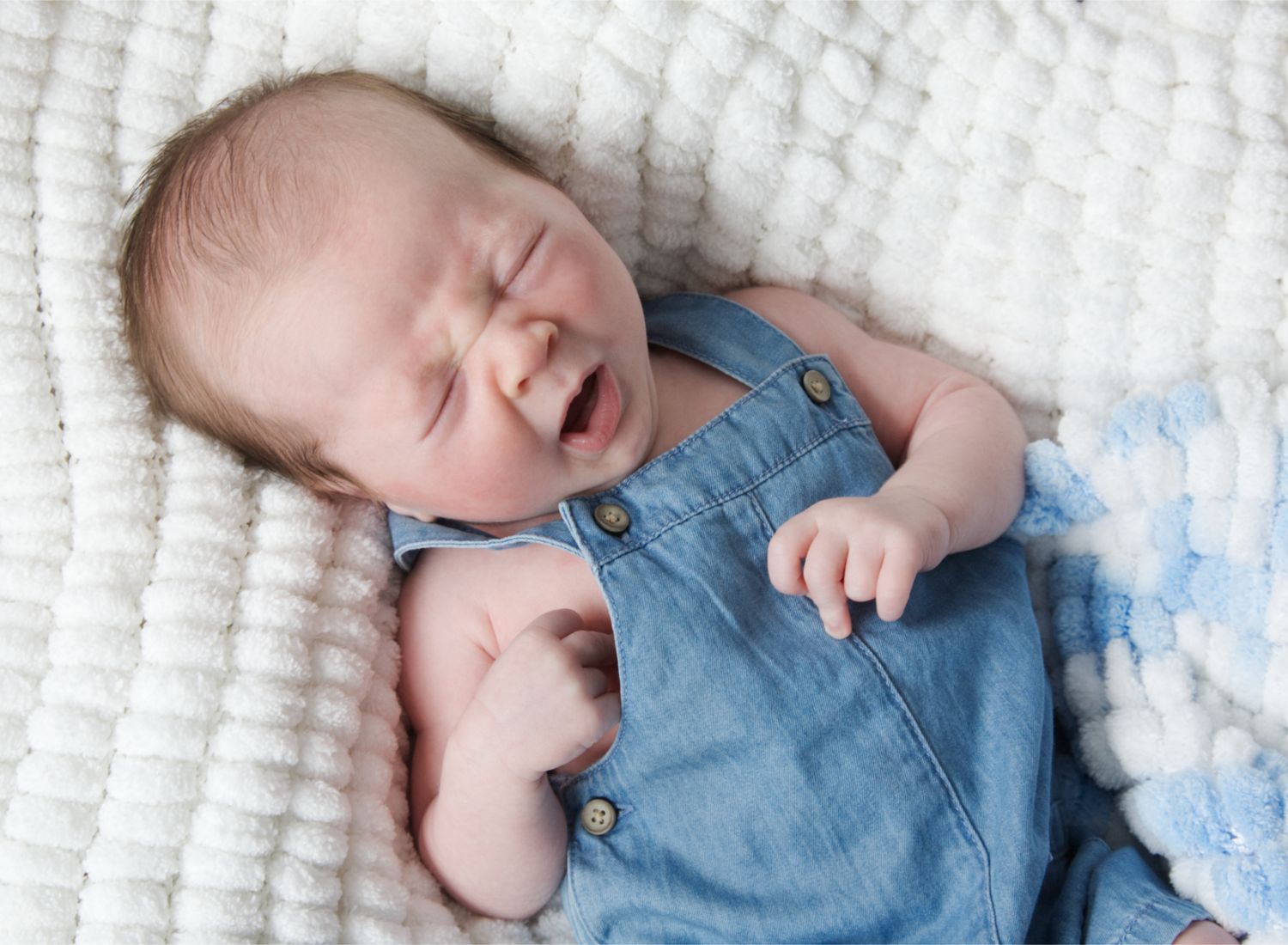 A yawning newborn wearing denim overalls lies on a white textured blanket.