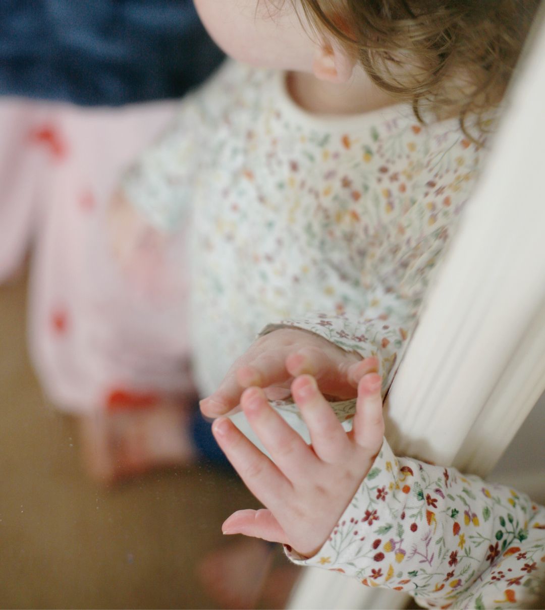 A child wearing a delicate floral top lightly touching a mirror with their small hands