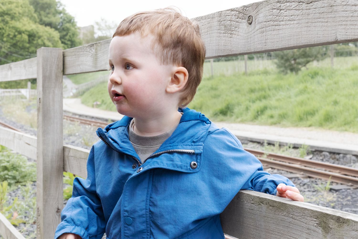 Child in blue raincoat sitting near wooden railway fence on cloudy day.