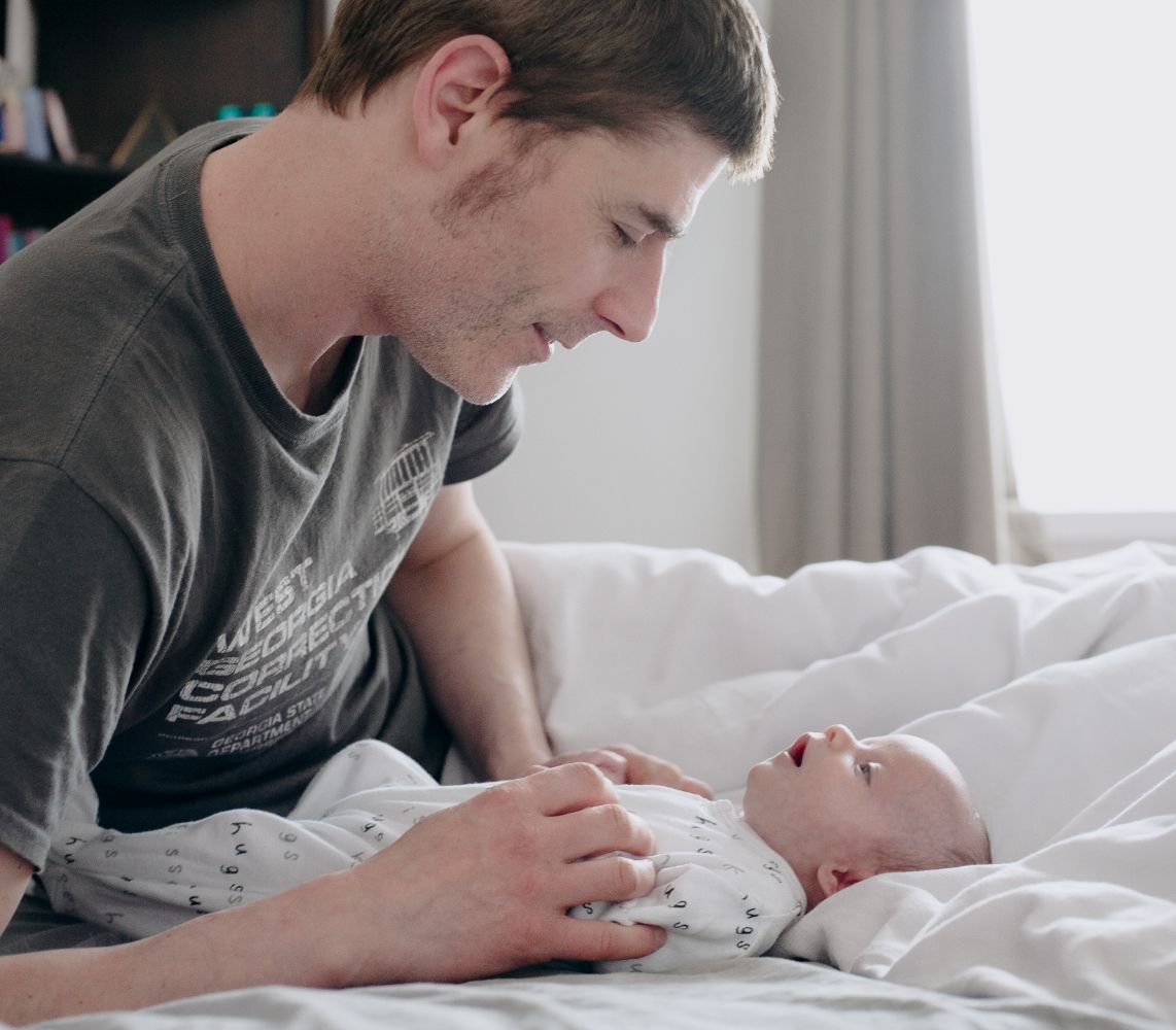 At-home photograph showing tender interaction between parent and newborn baby on soft white bedding.