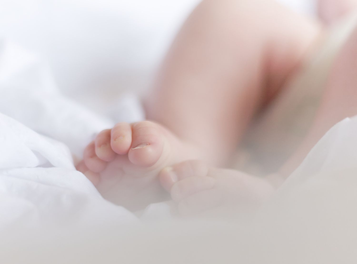 Tiny baby foot with delicate toes resting on soft white fabric in gentle, dreamy lighting.