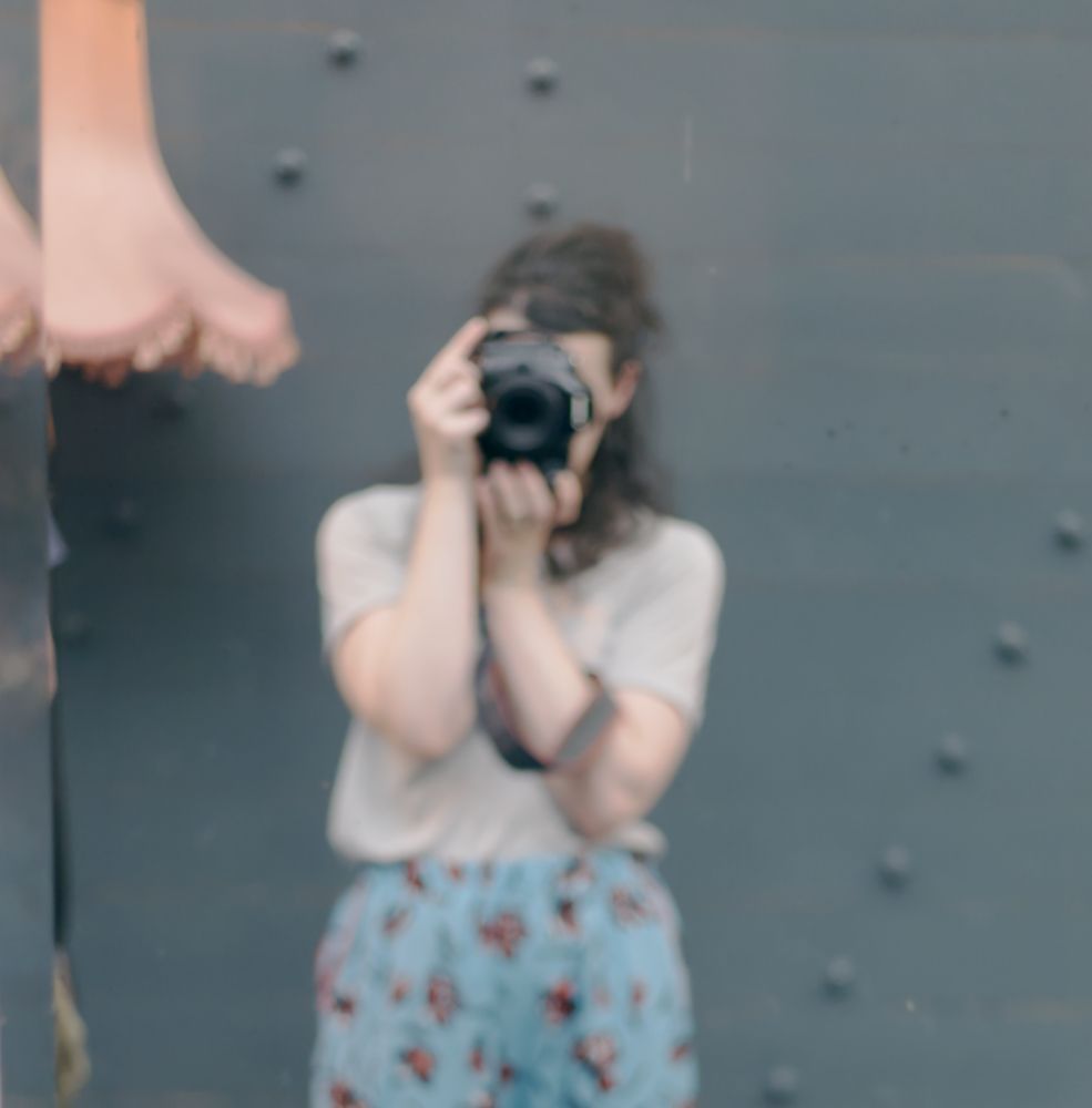 Blurred reflection of a photographer in a light sweater and floral skirt holding up a camera with a pink lamp in the background