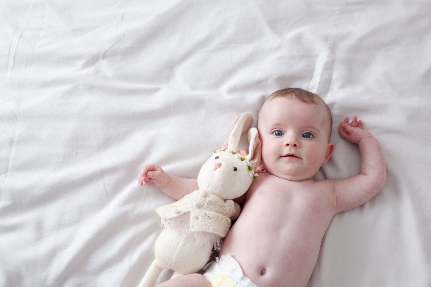 Baby photographed with soft natural light on white bedding with a handmade plush rabbit toy beside them