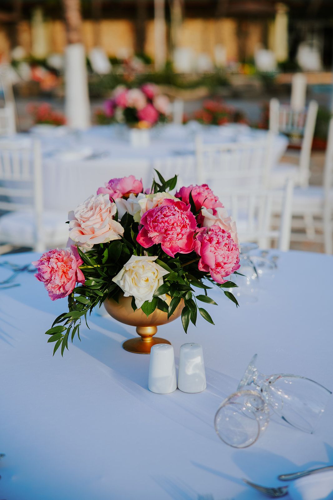 Pink and white peonies arranged in a gold vase on a white tablecloth at an outdoor wedding reception.