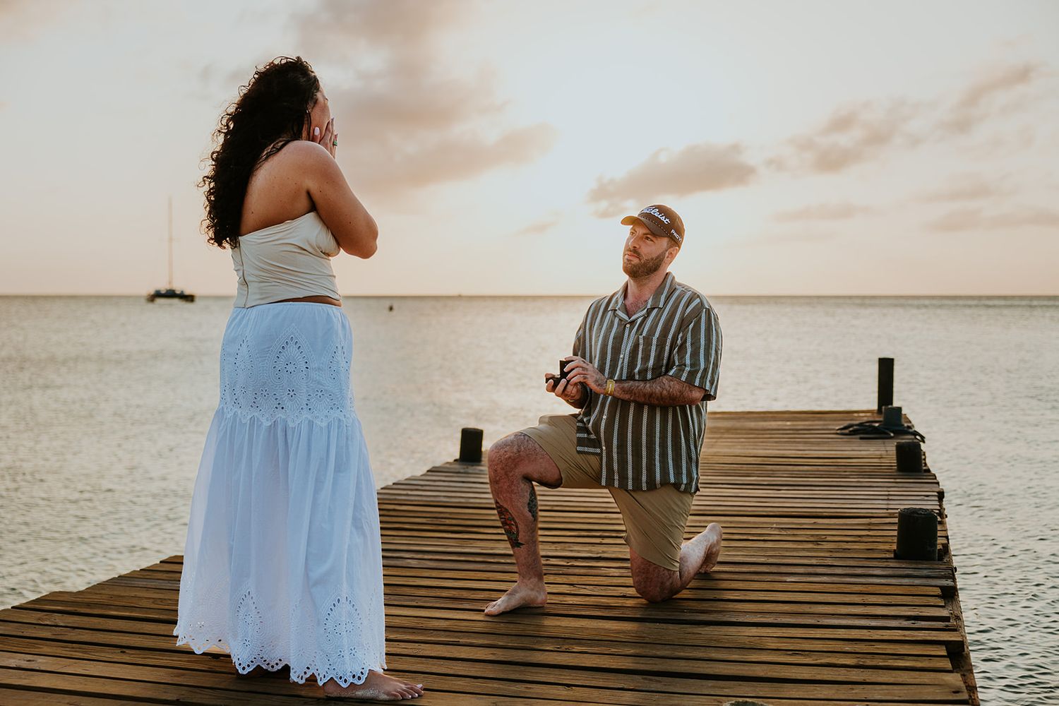 A couple sits together on a wooden dock at sunset overlooking a tranquil lake.