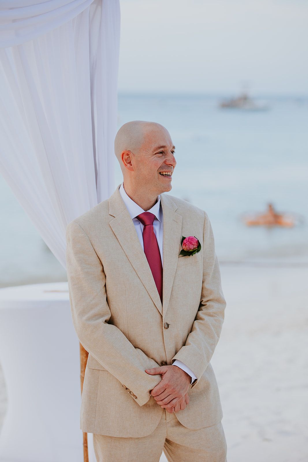 Groom in beige suit and red tie smiles on sunny beach with white curtains and ocean in background.