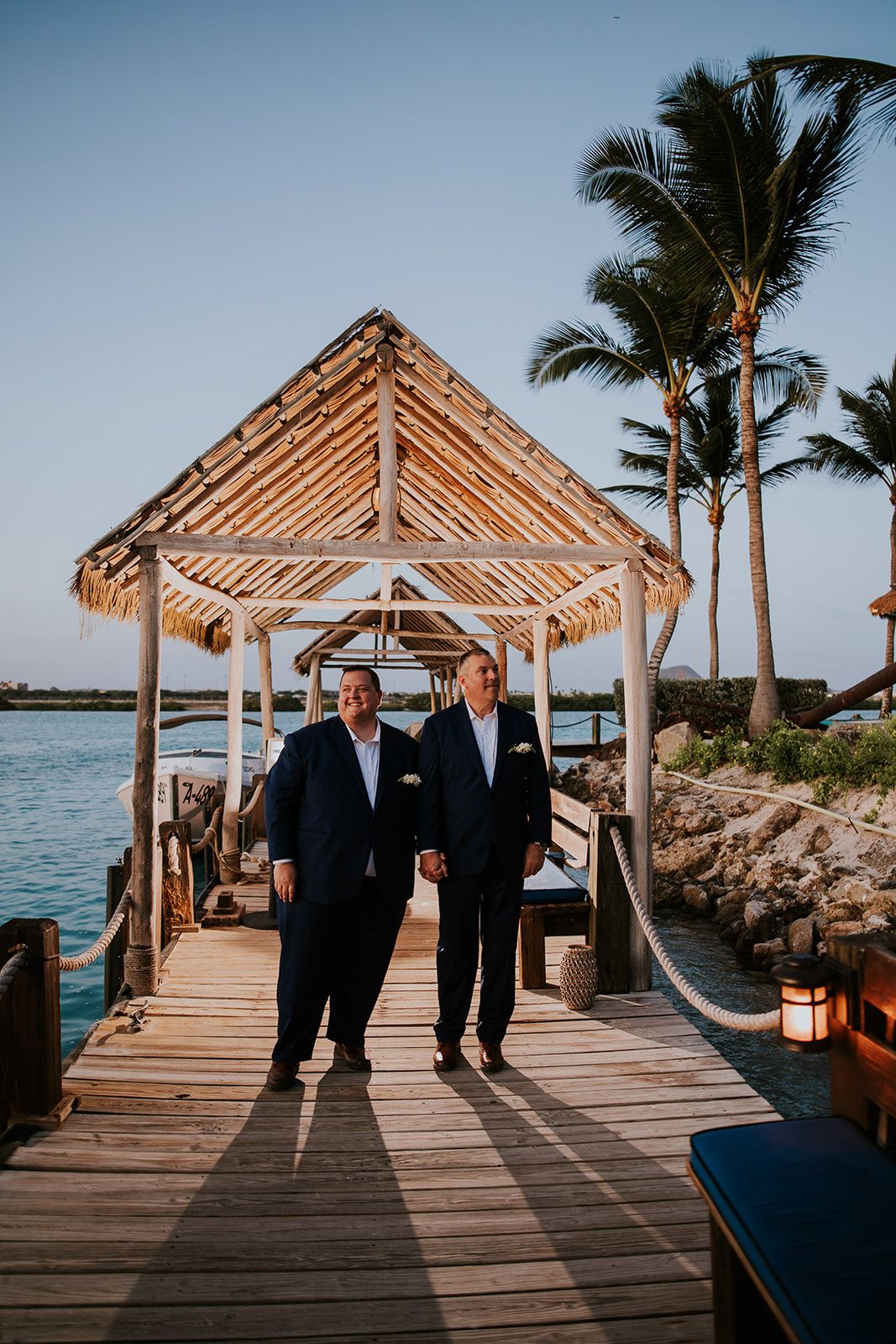 Two people walking along wooden pier with thatched gazebo and palm trees.