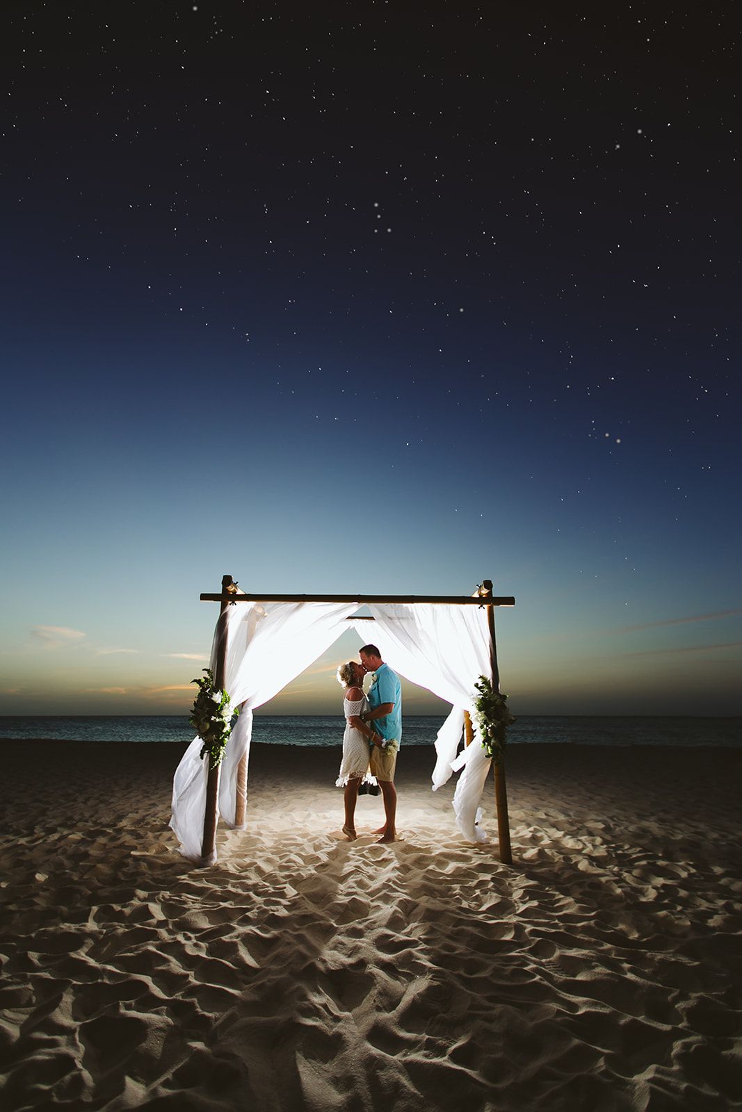 A romantic beach wedding ceremony setup with a bamboo arch draped in white fabric glowing under a starry night sky.