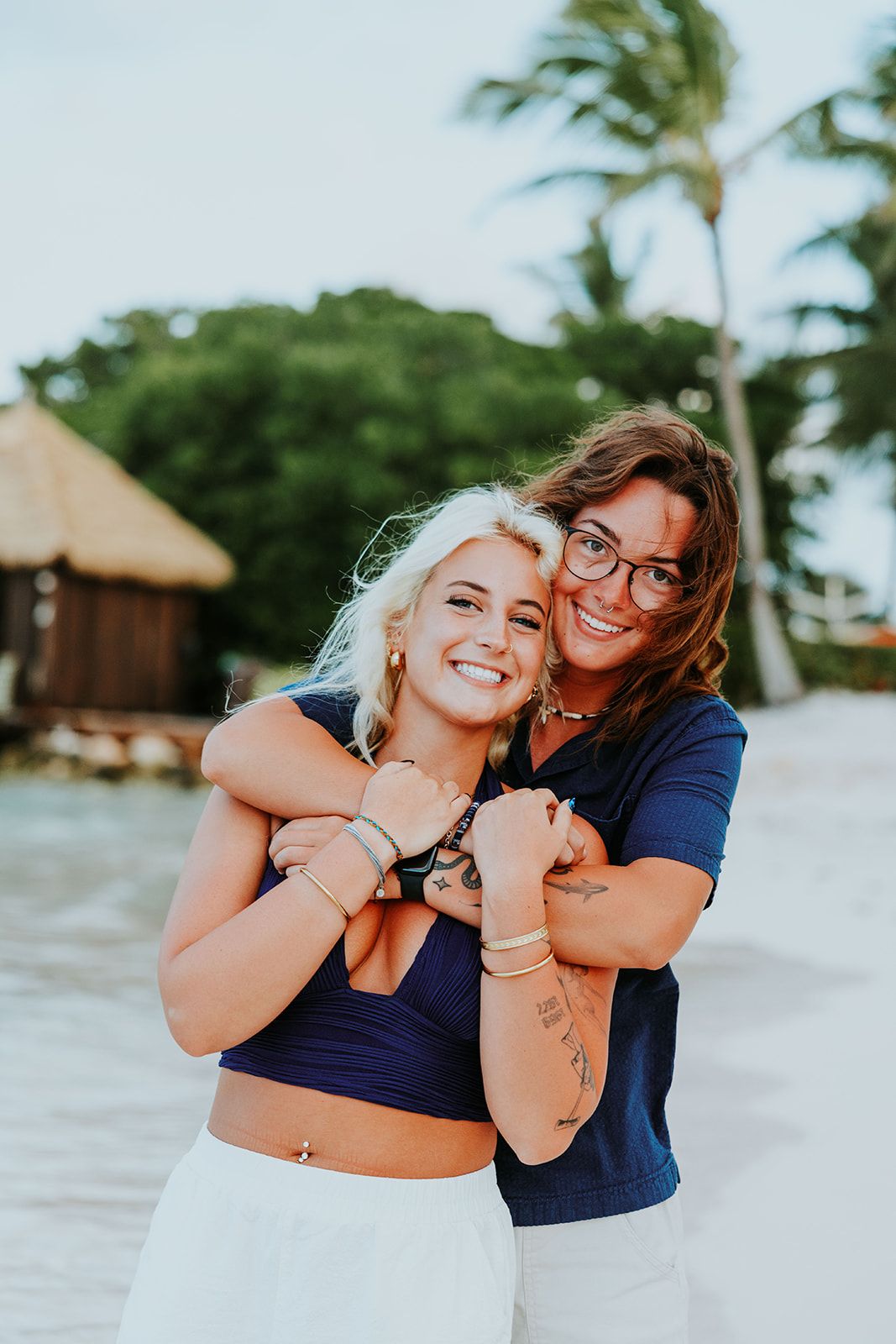 Two people share joyful embraces and laughter on a tropical beach with palm trees and thatched huts in the background.