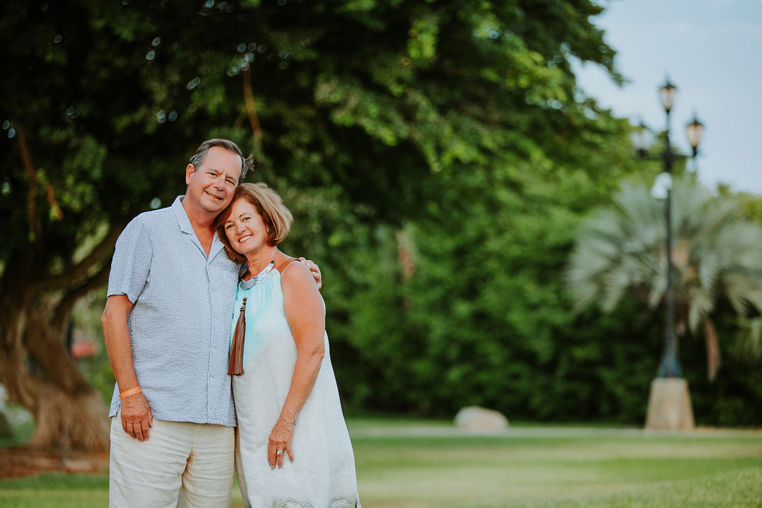 A couple in white summer attire embraces while standing in a lush green garden setting during golden hour.
