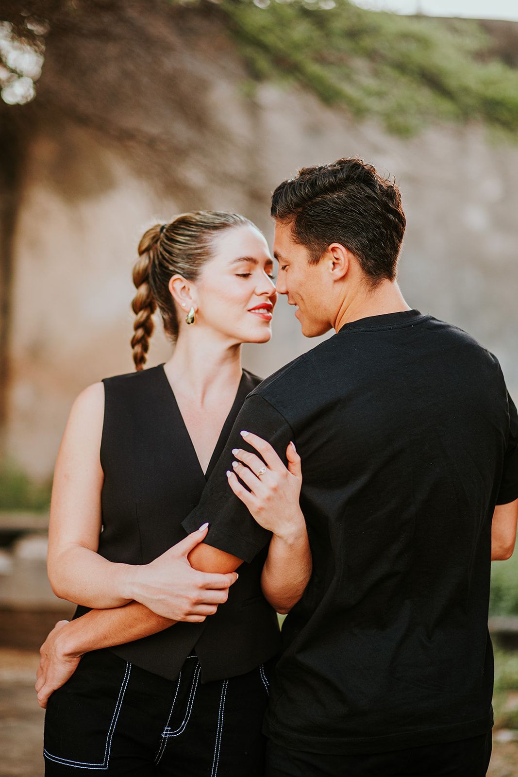 Intimate couple in black attire share romantic moment against natural backdrop.