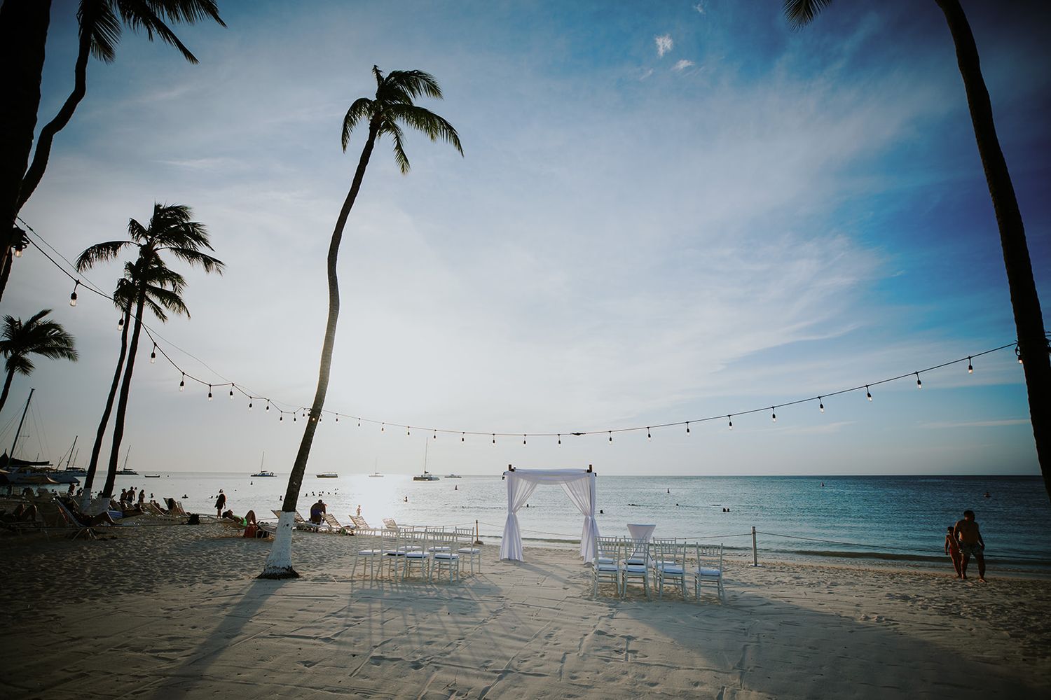 String lights hang between palm trees on a tropical beach at sunset with the ocean and blue sky in the background.