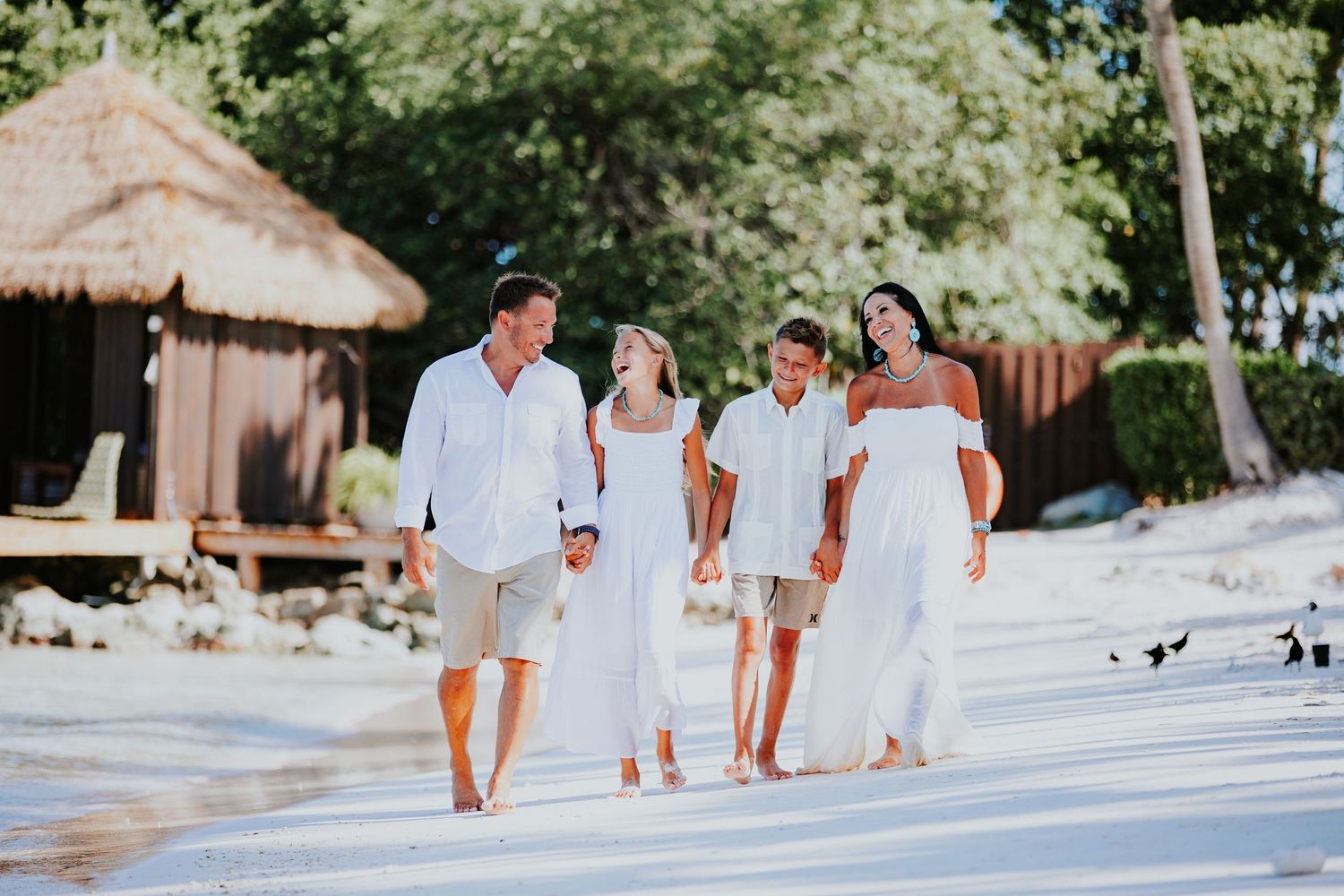 Family walks together in light colored clothing along a white sandy tropical beach.